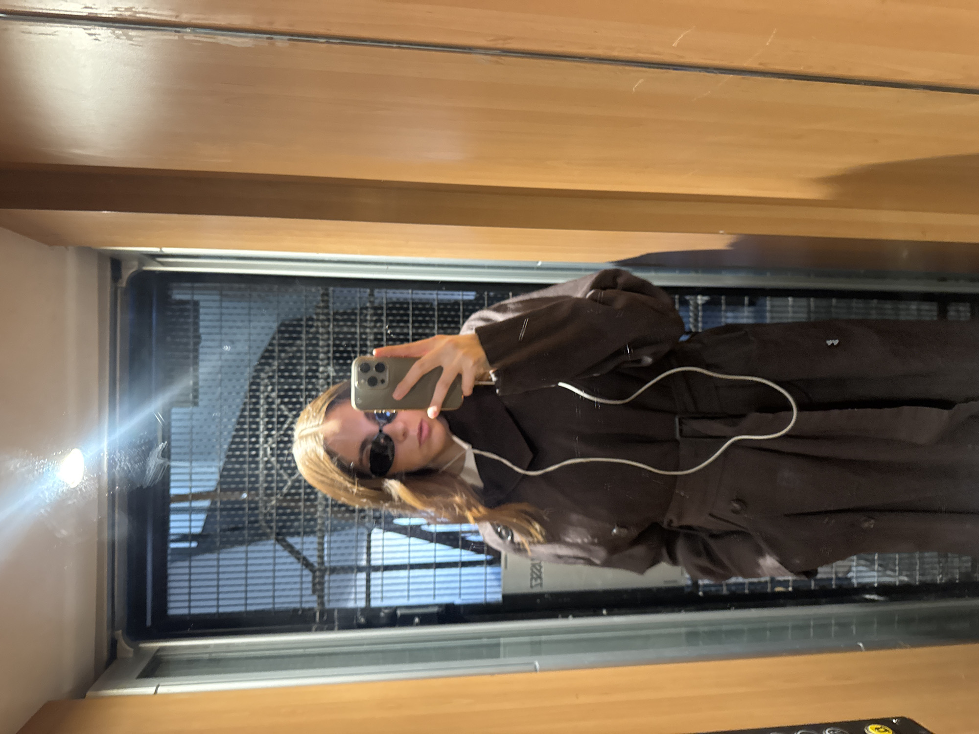 A woman is taking a selfie in an elevator with a grilled metal elevator shaft door. The woman is wearing sunglasses and a brown coat. She is holding a cell phone in her hand, and the flash is reflecting off the elevator door. The elevator's interior walls are a light wood finish. The buttons of the elevator are visible. The location is likely Paris, France, given the style of the elevator.