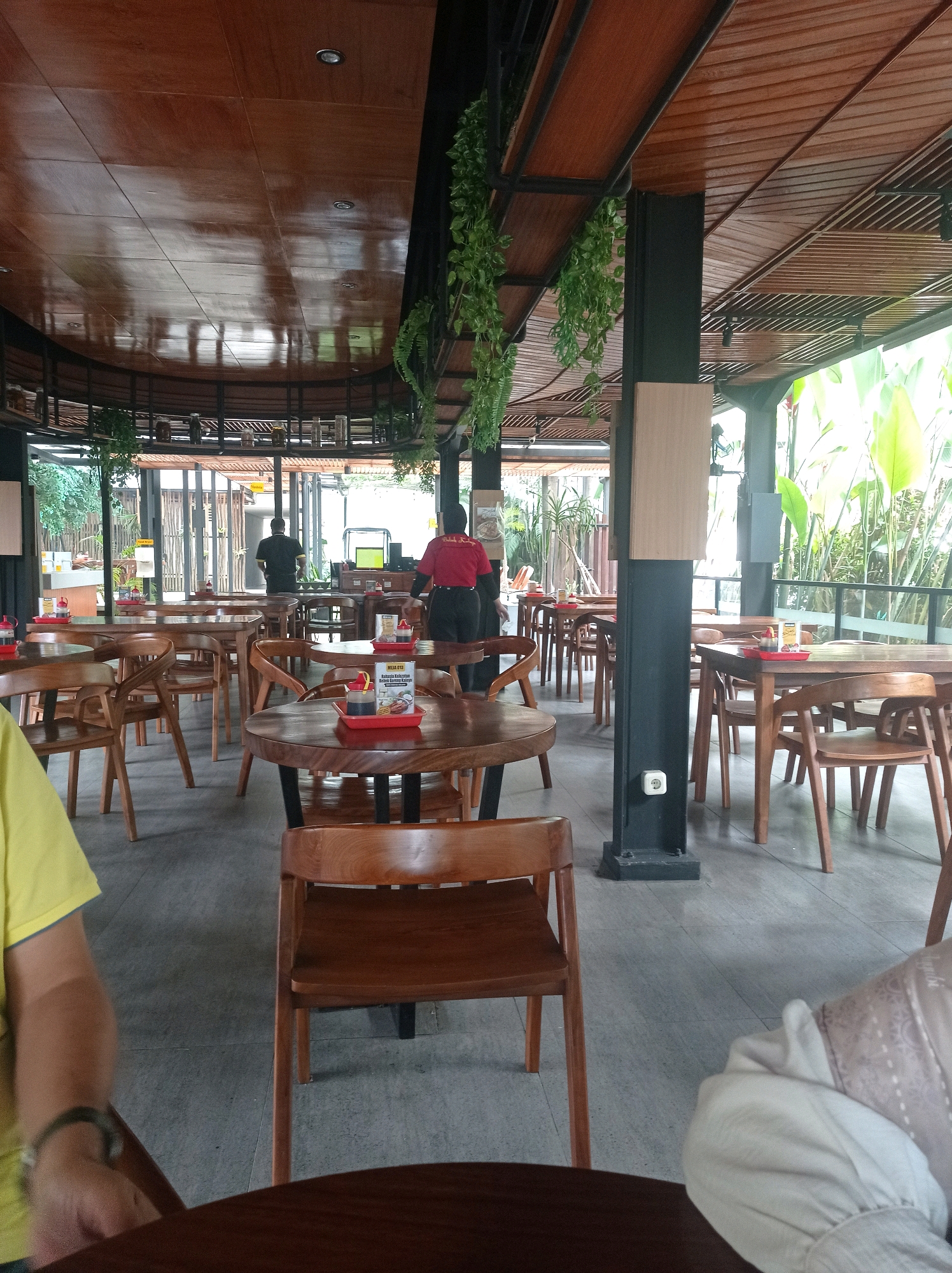 The image depicts the interior of a restaurant or cafe with a contemporary design, featuring wooden furniture and a slatted wooden ceiling. Several wooden tables and chairs are arranged in the dining area. In the background, two individuals are visible. One person, wearing a red shirt, appears to be a staff member, possibly a server, standing near a table. Another person, wearing a dark shirt, is also present. Hanging plants add a touch of greenery to the space, and large windows allow natural light to filter in, revealing lush vegetation outside. The floor is tiled in a light gray. The scene suggests a casual dining environment.