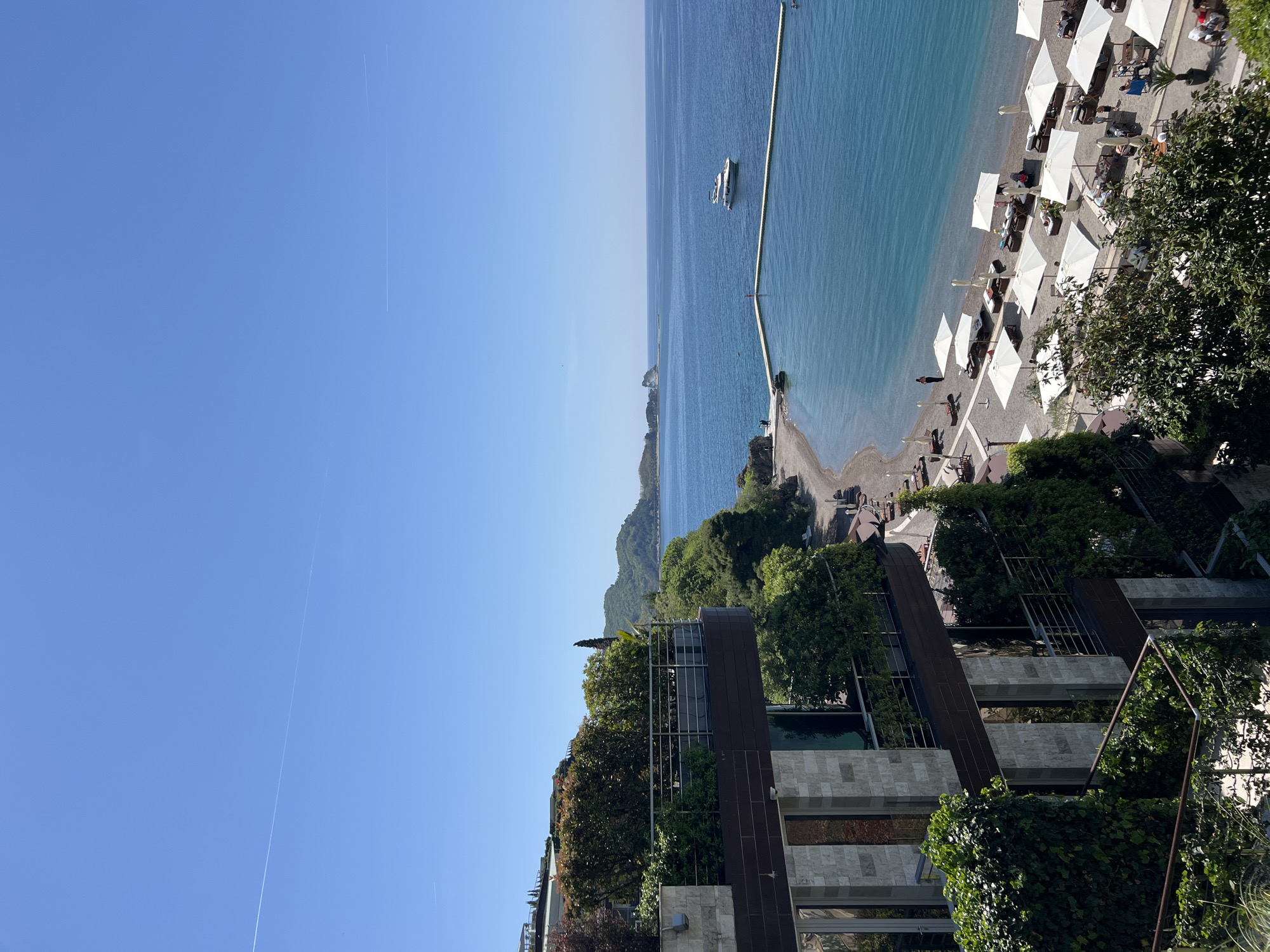 This panoramic image, likely taken from an elevated perspective in Budva, Montenegro, captures a serene coastal scene under a clear blue sky.
The upper portion of the frame is dominated by a vast, unblemished blue sky, with a faint, thin contrail visible from an aircraft. Below the horizon, the calm, blue-green waters of the Adriatic Sea stretch out, reflecting the sky's brightness. A long, light-colored breakwater or pier extends significantly into the water, with a white yacht or boat anchored nearby, adding a touch of leisure to the seascape. In the distance, undulating green hills and mountains rise from the coastline.
The foreground and mid-ground reveal a well-appointed beach area and modern resort architecture. A pebble beach is lined with numerous white, open beach umbrellas providing shade, accompanied by sun loungers. Several people can be seen relaxing on the beach, some near the water's edge, enjoying the sunny weather.
Adjacent to the beach, and extending upwards into the lower-left of the frame, are contemporary multi-story buildings, possibly part of a hotel or luxury complex. These structures feature a blend of materials like dark metal, stone, and large glass windows, and are extensively integrated with lush green vegetation, including trees, shrubs, and climbing ivy covering facades and balconies, creating a verdant and inviting environment.
The time of day appears to be mid-morning to early afternoon, indicated by the bright, clear sunlight and lack of harsh shadows. The overall atmosphere is one of peaceful relaxation and natural beauty typical of a Mediterranean resort destination. No discernible text is visible in the image.