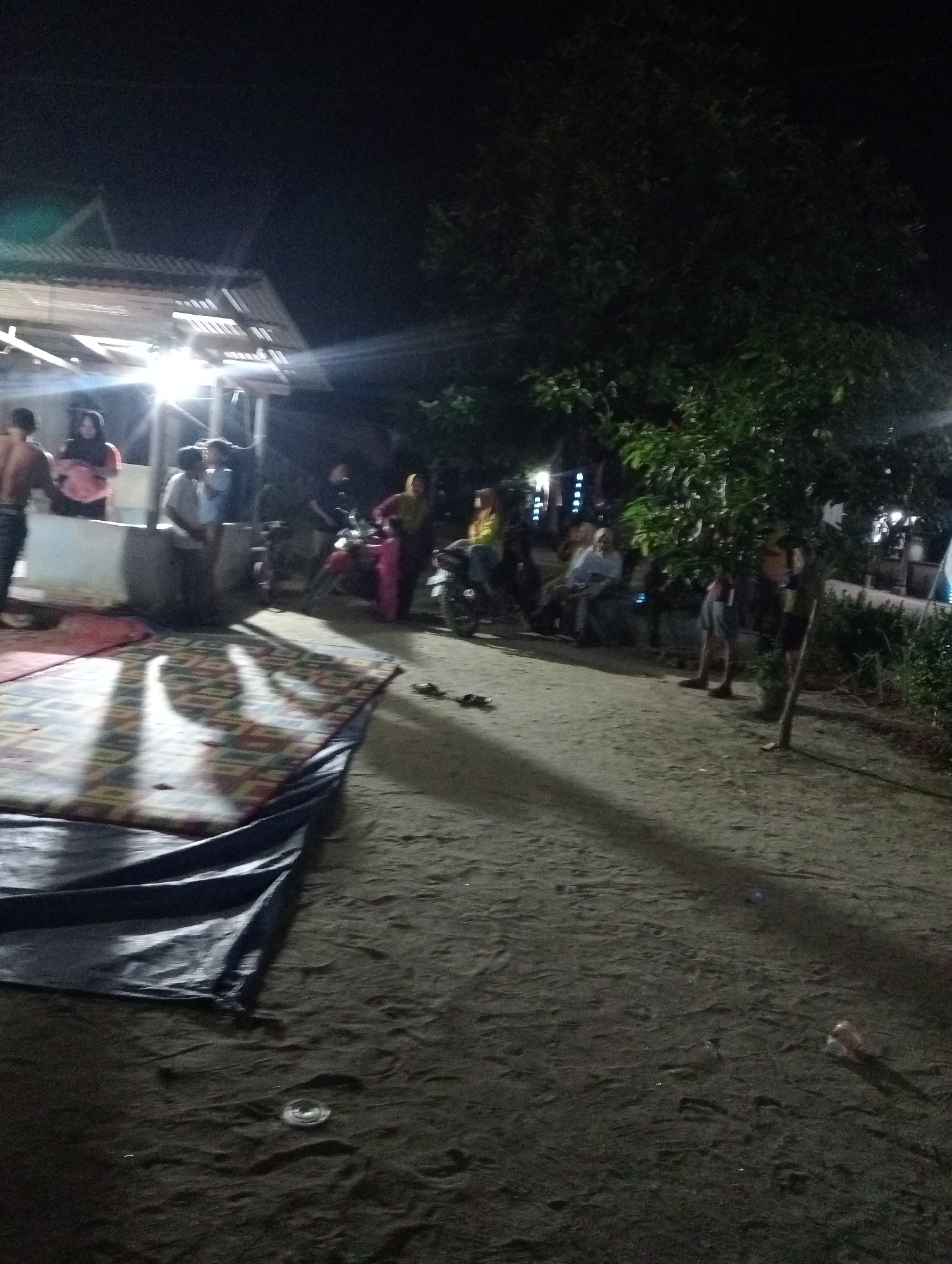 The image captures an outdoor scene at night, likely in Rasuan Baru, Indonesia. The lighting suggests artificial illumination, possibly from a nearby structure, casting strong shadows. In the background, a building with a corrugated roof is visible, with a bright light source emanating from it. A group of people are gathered near this structure, some standing and others appearing to be seated. Two motorcycles are parked in the vicinity, and one person is near a pink-colored garment.
In the foreground, a large mat or blanket is spread on the ground, with a dark tarp partially covering it. The ground itself appears to be sandy or dirt. The overall impression is of a casual gathering or event taking place in a somewhat rustic outdoor setting. The presence of multiple individuals, vehicles, and a mat suggests a social or community activity. The darkness of the night is punctuated by the artificial lights, creating a dynamic interplay of light and shadow that obscures some finer details.