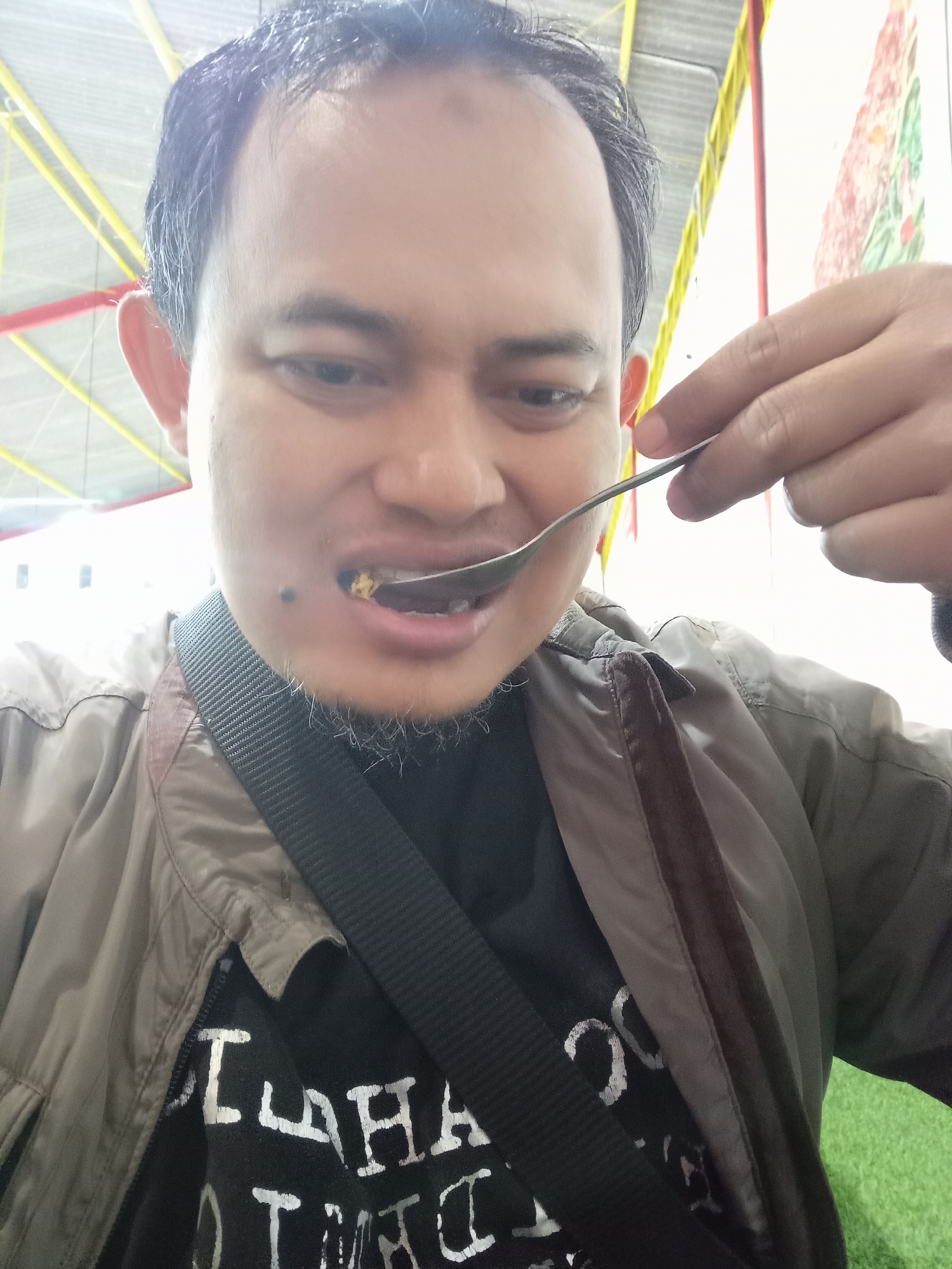 A man with short, dark hair and a light beard is taking a selfie while eating. He is wearing a brown jacket over a black t-shirt with white text. A black strap runs across his chest. He is holding a spoon with food up to his mouth. The background appears to be an outdoor covered area with yellow and red supports visible. The scene is likely in Landungsari, Indonesia.