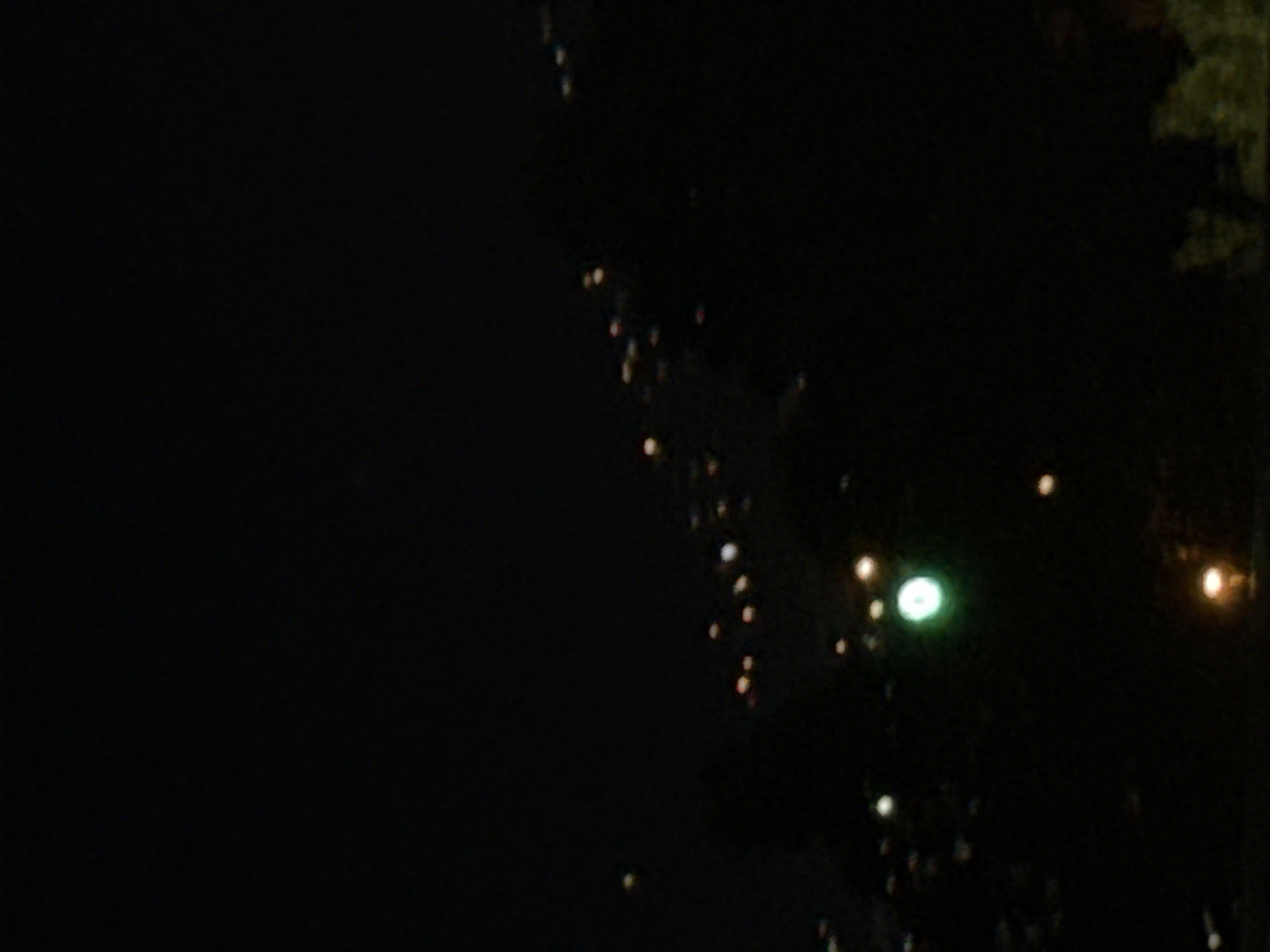 A nighttime, out-of-focus aerial view shows blurred city lights in Tbilisi, Georgia. The image captures distant, indistinct lights scattered across a dark, hilly landscape, suggesting a city at night. The lights vary in intensity and color, with some appearing as small, white or yellow orbs, while others are larger and more diffused, including a prominent green light. The overall impression is that of a sprawling urban area illuminated against the darkness of the night.