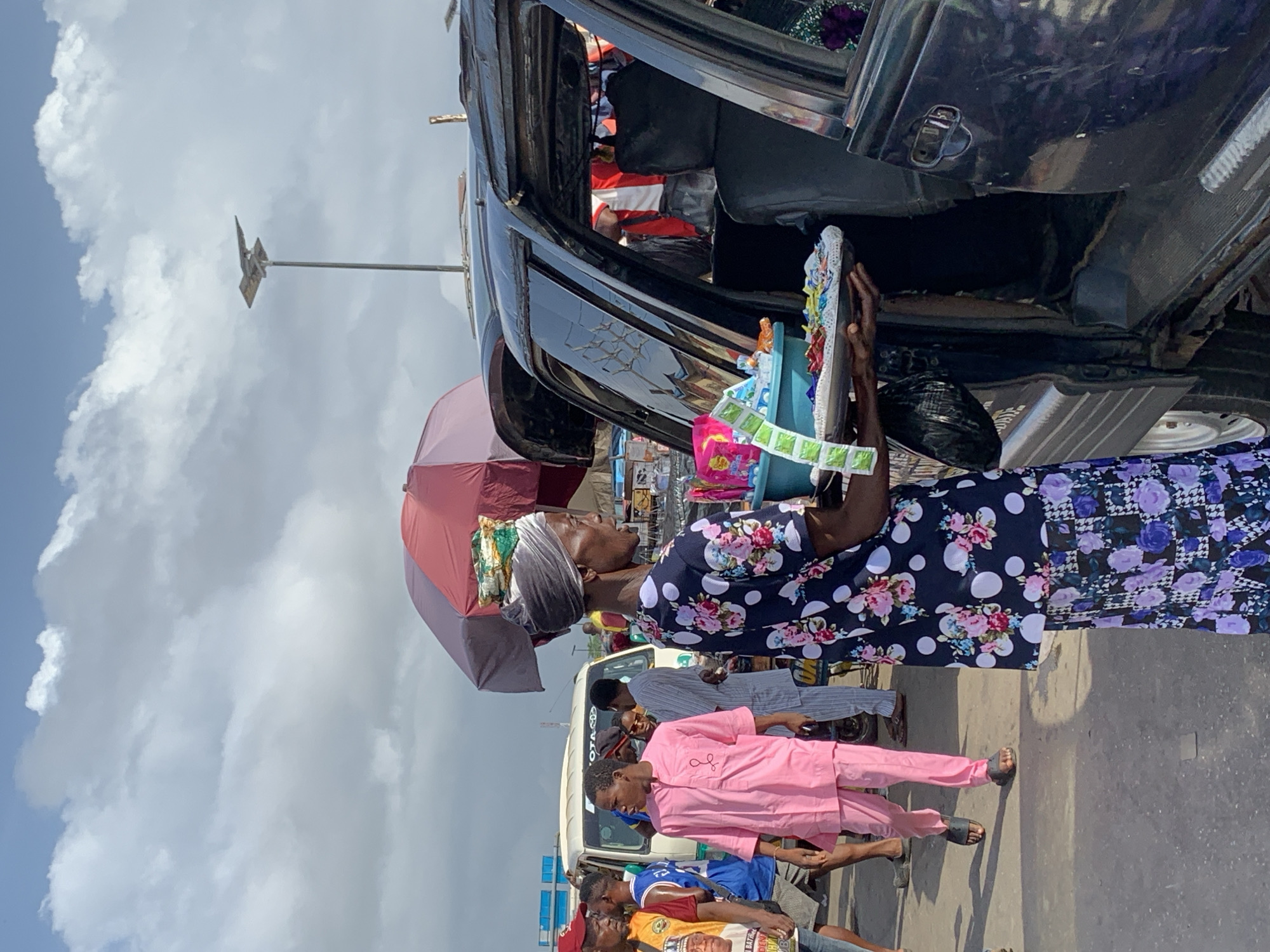 A street vendor stands in Ikorodu, Nigeria, selling goods to passengers inside a vehicle. She holds a white tray filled with small items, possibly candies or snacks. The vendor uses a maroon umbrella to shield herself from the sun. She is wearing a dress with floral prints. Several pedestrians are nearby. The setting is an urban environment with a cloudy sky.