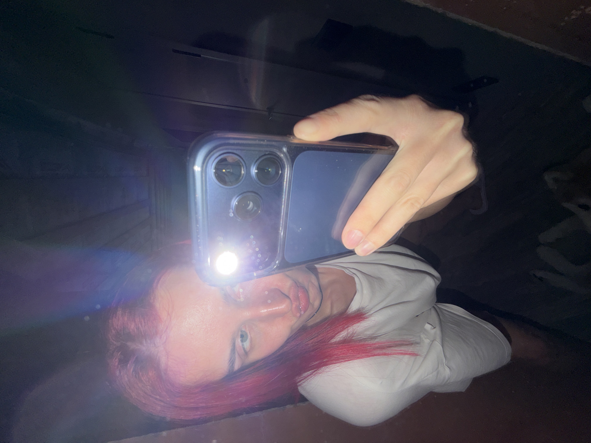 A top-down, dimly lit photograph captures a young individual taking a selfie or self-portrait in a very dark environment. The subject has vibrant red hair, fair skin, and striking blue eyes, which are directed towards the camera. They are wearing a white t-shirt and have several small, white facial piercings around their mouth and possibly nose.
The photograph is taken with a smartphone, likely an iPhone due to its triple camera array and clear case, which is held in the subject's hand. The phone's flashlight is actively on, serving as the sole light source and creating a bright glare and lens flare that partially obscures the upper part of the person's face.
The background is uniformly dark and indistinct, offering no specific details about the setting beyond it being a low-light space, suggesting nighttime or an interior with no external light. The strong illumination from the phone highlights the person's features and hair against the dark backdrop. No visible text is present in the image. The location context provided is Chaikovsky, Russia.