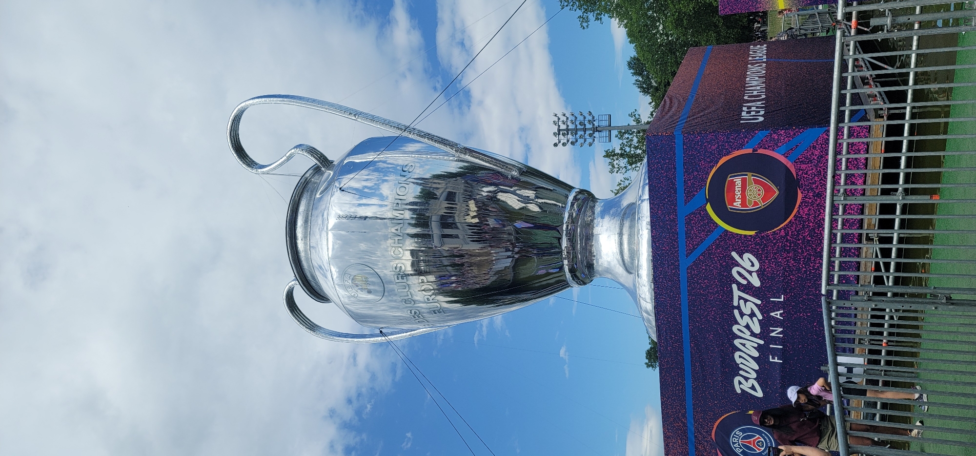 The image shows a large, reflective replica of the UEFA Champions League trophy, suspended in the air via cables against a backdrop of a blue sky with scattered clouds. The trophy's surface reflects the surrounding environment. A promotional banner is visible to the right, displaying team logos, including Arsenal, and the text "Budapest 26 Final," indicating an association with the UEFA Champions League Final potentially hosted in Budapest. A metal fence is positioned in the foreground, obscuring a partial view of people. The event seems to be held outdoors. The presence of promotional material and people suggests a public viewing or fan zone related to the UEFA Champions League. The location is assumed to be in Budapest, Hungary.