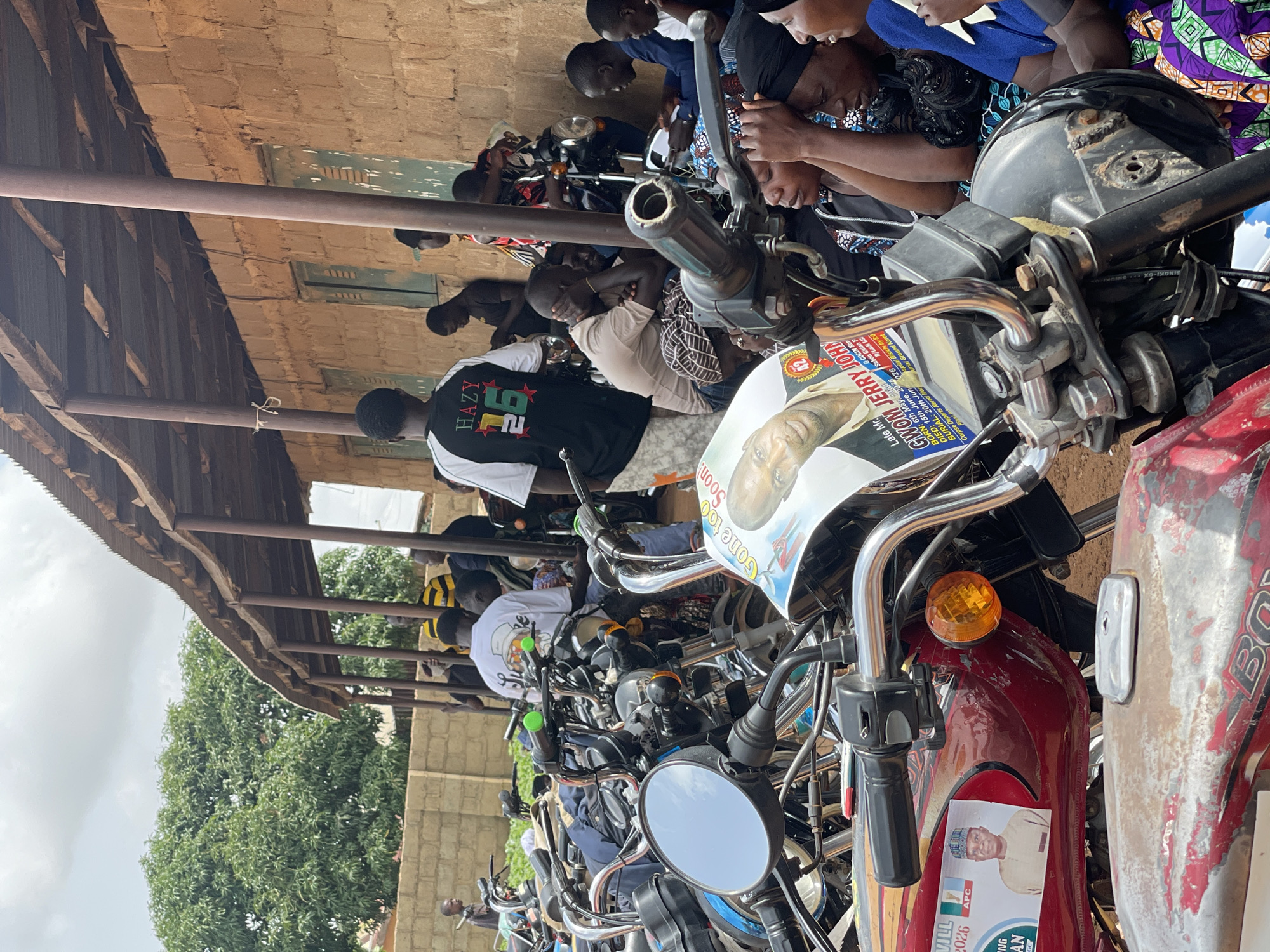 The image depicts an outdoor scene in Kassa, Nigeria, characterized by a gathering of people and numerous motorcycles parked under a covered structure. The structure appears to be a rudimentary building with a corrugated metal roof supported by wooden poles, adjacent to a brick wall. A large tree with dense green foliage is visible in the background.
In the foreground, several motorcycles are prominently displayed, some with visible political campaign materials. One motorcycle has a sticker featuring a man's portrait and text that reads "Late Mr. GWOM JERRY JOHY". Another motorcycle displays a sticker with the image of a man in traditional attire and the text "APC".
A group of individuals, predominantly men, are seen interacting and observing. Some are standing, while others are seated or leaning against the motorcycles. Their attire varies, with some wearing casual clothing and others in more formal or traditional garments. The mood appears to be somber or contemplative.
The environment suggests a local community setting, possibly a gathering for a political event or a community meeting. The presence of so many motorcycles indicates that transportation is a key element of this gathering, likely involving people from the surrounding area. The overall scene conveys a sense of community and political engagement within Kassa, Nigeria.