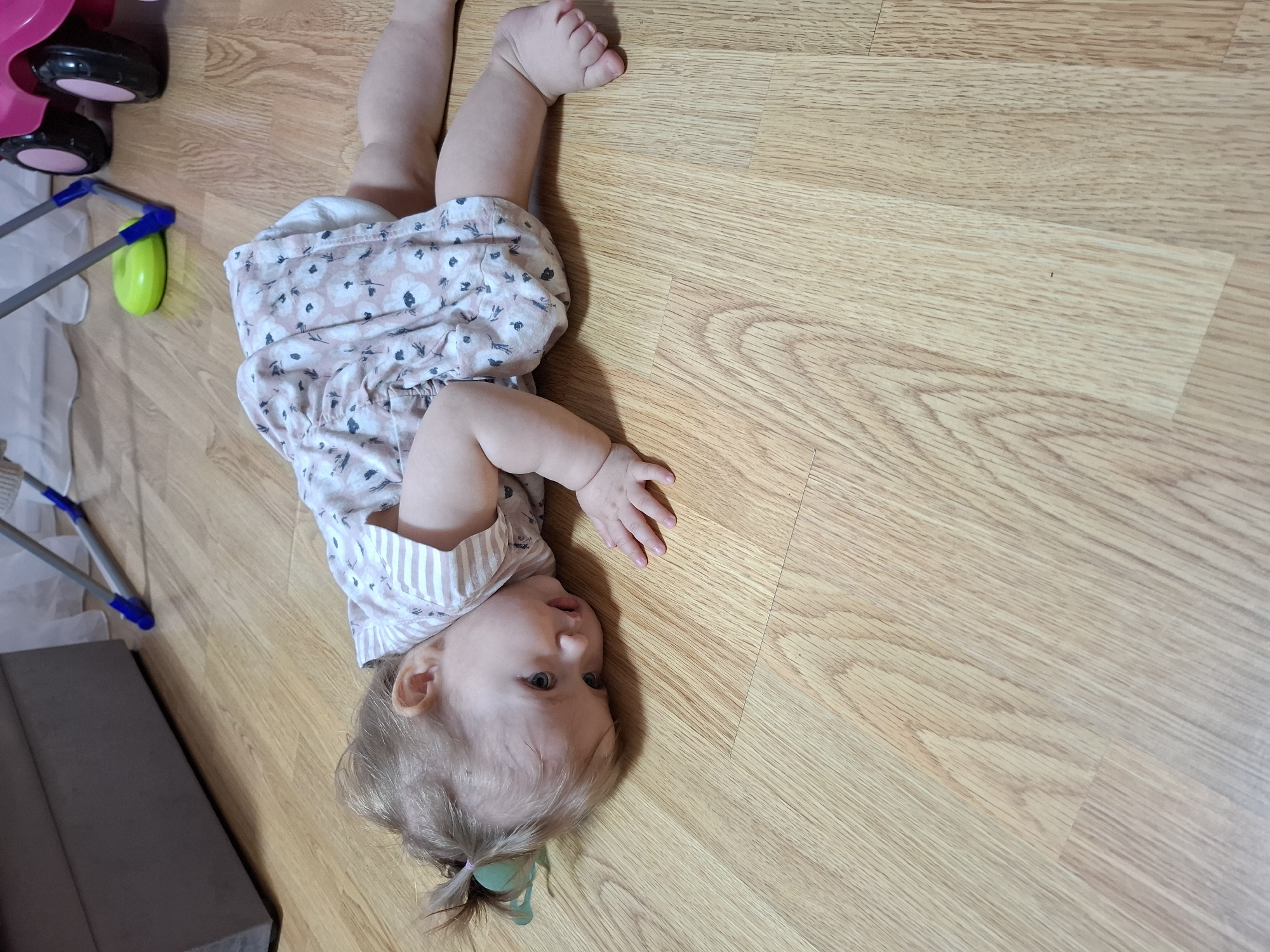 The image depicts a baby lying on a light wood-grain floor. The baby is wearing a light pink and white patterned romper. Their arms are outstretched. The baby's hair is blonde and pulled back into a small ponytail. In the background, there is a pink and black toy and a metal and plastic stroller-like object.
While the location might be Chaikovsky, Russia, I don't see any evidence of that in the picture.