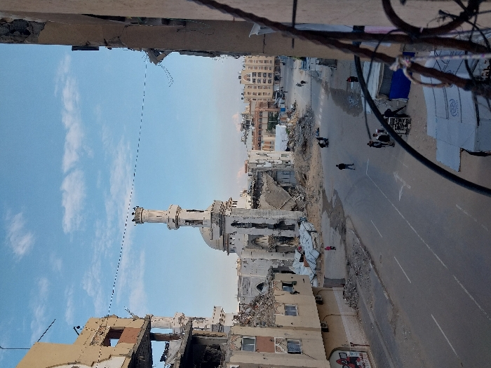 A war-torn city street scene features significant destruction. A mosque stands amidst the rubble, its minaret still intact. Buildings show extensive damage, with gaping holes and crumbled facades. People walk on the road near tents, possibly for shelter. Debris and rubble are scattered everywhere. The sky is light blue with scattered clouds. Overhanging cables and part of a building frame the upper part of the shot.