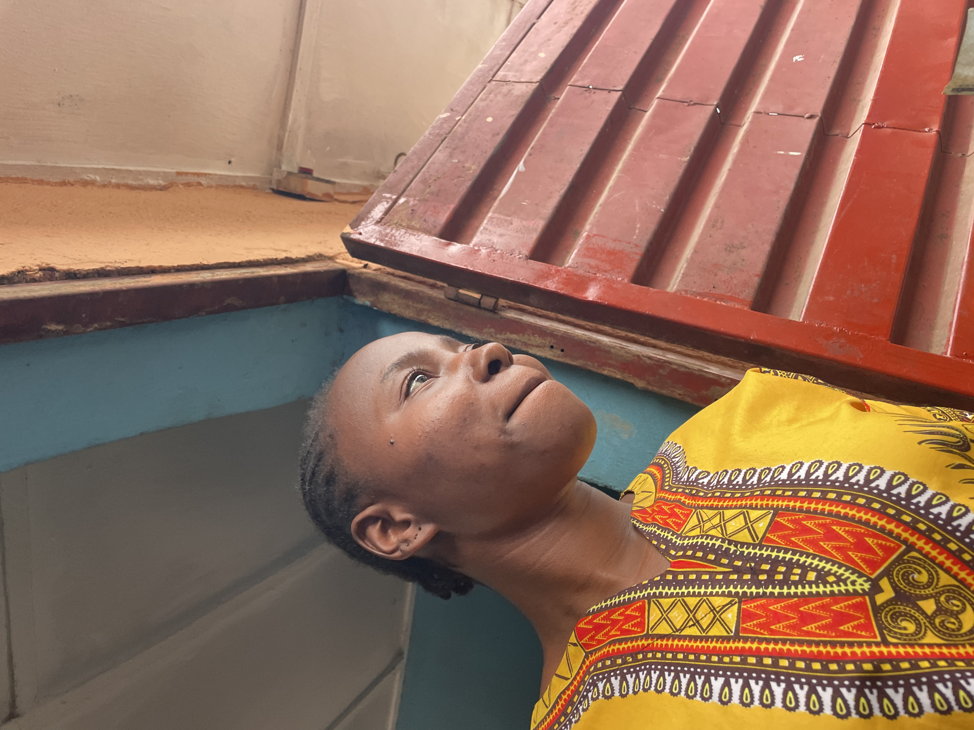 A low-angle shot shows a woman with dark skin looking upward. She wears a yellow and red patterned top. Above her is a rusty red metal lid or cover. The background includes light blue painted walls and a beige-painted surface. The location is not explicitly discernible as being Jos, Nigeria from visual evidence.