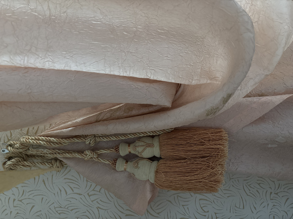 This image depicts an interior scene in the city of Velikiye Luki, Russia. A light pink curtain, characterized by a visible crushed or crinkled texture and subtle pale gold, possibly embroidered, leafy patterns, is draped and drawn to the left. It is held open by a decorative tie-back made of braided light gold rope. The rope forms two knots and is secured to a metallic, silver-toned wall hook located at the upper left. Hanging from the rope are two identical tassels, each featuring a contoured, gourd-shaped head wrapped in light beige cord with distinct peach-toned raised piping. Below each head, a voluminous fringe of fine, medium peach-brown strands extends downwards. To the left of the curtain and tie-back, a light-colored wall is visible, adorned with wallpaper that displays a repeating pattern of stylized, organic shapes in a subtle gold hue. The scene is illuminated by soft, diffused light, highlighting the textures of the fabric and wall covering. No individuals are present, and no specific actions are underway.