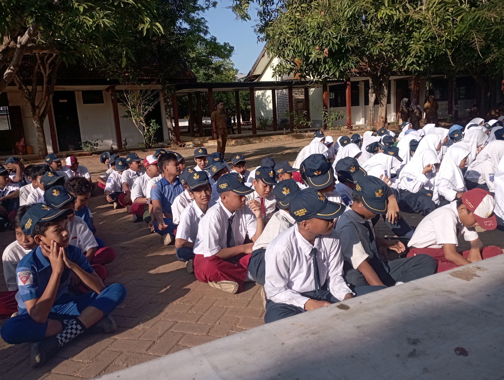 A group of students in uniforms and caps are sitting on a brick-paved ground in an outdoor area. They appear to be in front of a building with an open-air walkway. The students are facing in various directions, some looking towards the foreground and others looking away. Some students are wearing white shirts and dark pants or skirts, while others are in blue uniforms. The environment is sunny with trees and a building visible in the background. A man in a brown uniform is standing in the background. The scene suggests a school or educational setting.