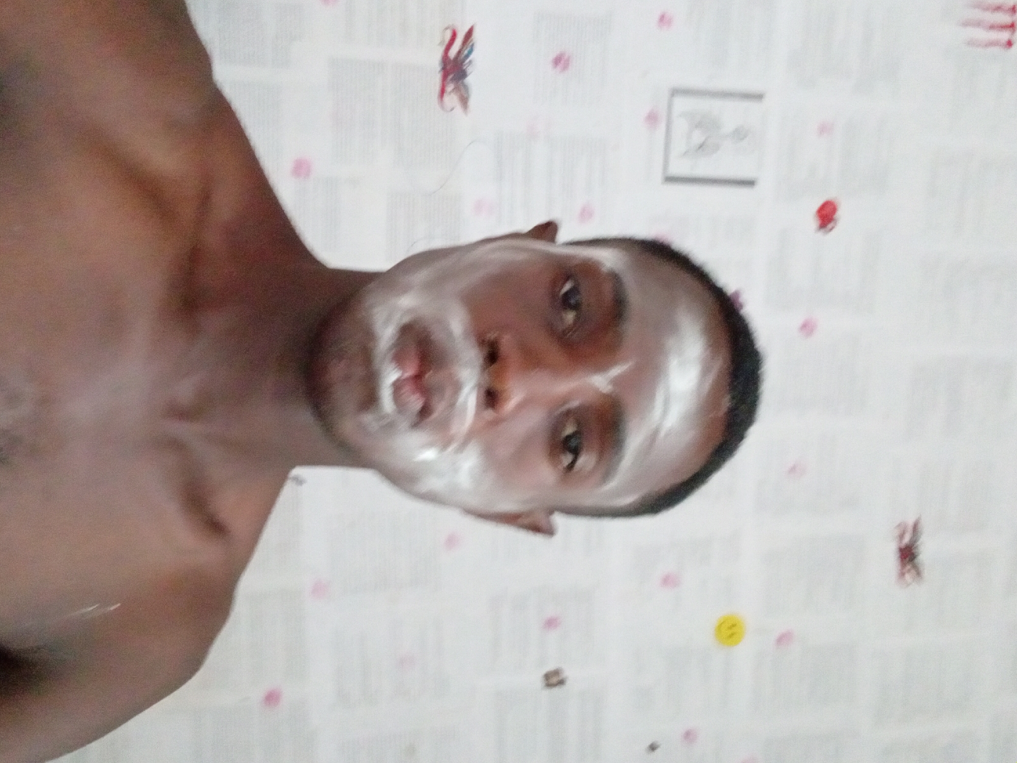 A portrait-style photo of a dark-skinned person with short dark hair, possibly male, with a white substance covering their face. The person is shirtless and facing the camera. The background consists of a patterned white surface with various small red and yellow designs scattered across it. The location, Akwete, Nigeria, cannot be confirmed based on the visual information.