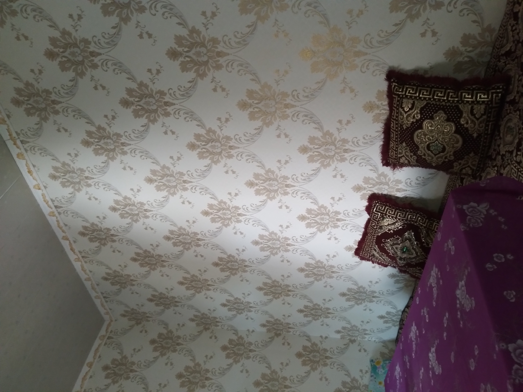 The image shows an interior corner of a room. The walls are covered in white wallpaper with a repeating damask pattern in beige and gold. The ceiling appears to be white or light gray. In the foreground, on the left, is a portion of a bed or sofa covered with a dark purple cloth patterned with white flowers. Two decorative pillows with dark red covers and gold embroidery are placed on the sofa. The room appears to be a residential interior. The location is described as Baki, Uzbekistan, which is a village in the Surxondaryo Region of Uzbekistan.