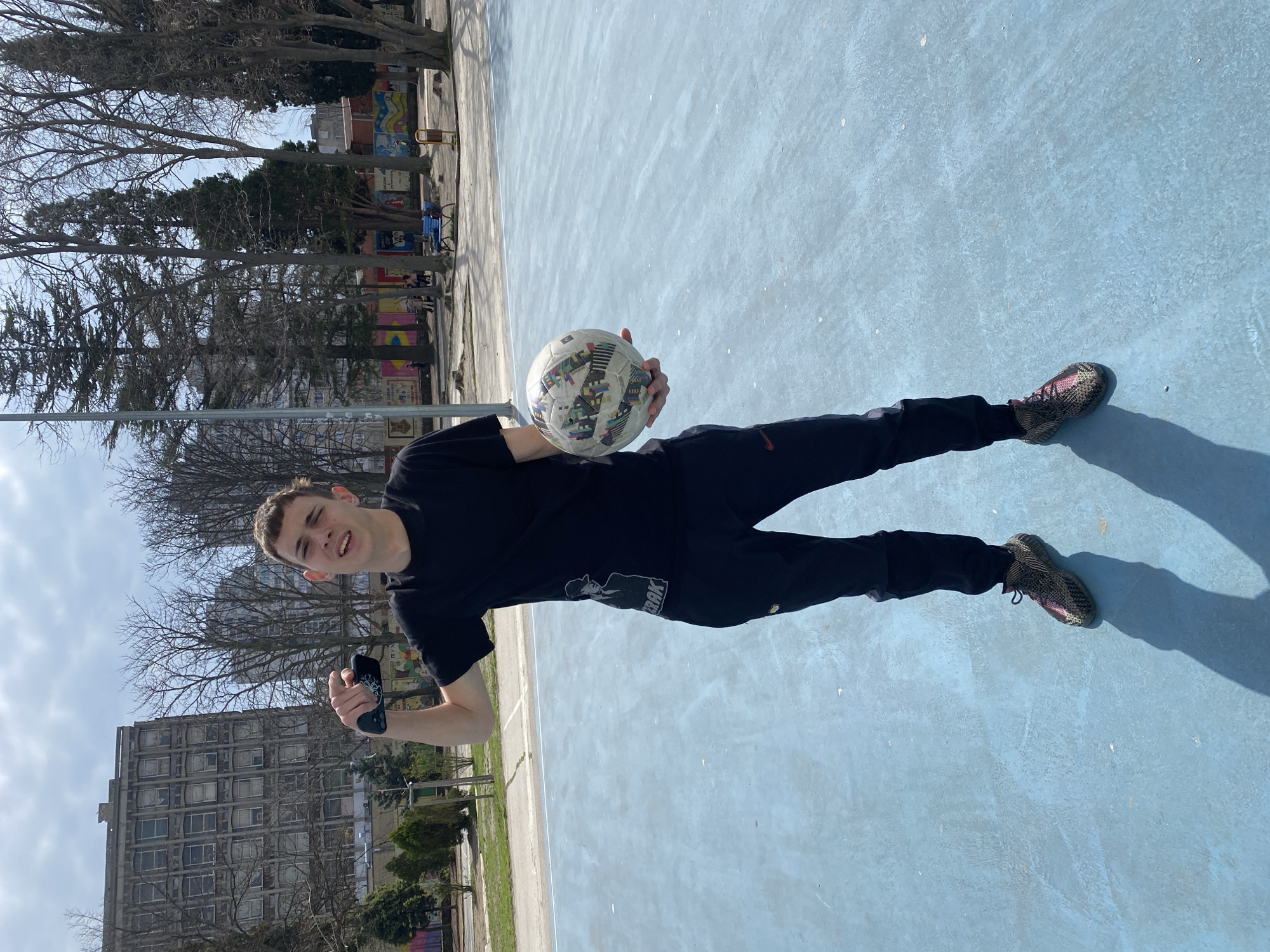 A young man in a black t-shirt and black sweatpants with a graphic on the leg stands on a light blue concrete surface, holding a soccer ball and a smartphone or game controller. He is looking at the camera and appears to be smiling slightly. The environment suggests an outdoor urban area, possibly a park or recreational space. Behind the man, there are bare trees, a tall, aged concrete building with many windows, and a mural on a low wall. Given the style of architecture and the general urban layout, it is likely that the photo was taken in Varna, Bulgaria.