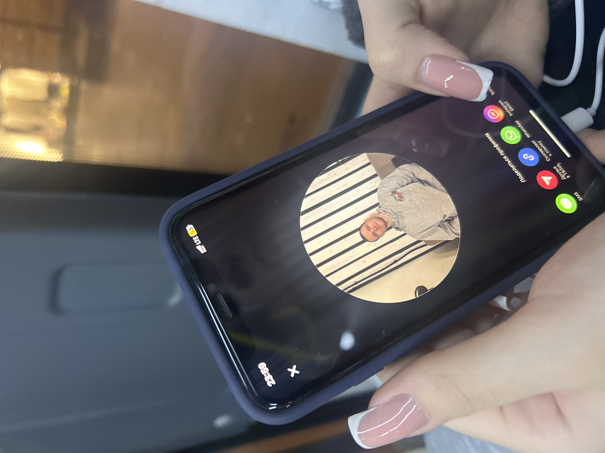 A close-up shot depicts hands holding a dark blue-cased smartphone indoors. The hands feature manicured nails, with some showing French tips and others a pink polish. A white charging cable is connected to the phone.
The smartphone screen displays a profile-sharing interface in Russian. A circular profile picture shows a young man wearing a grey hooded sweatshirt. Below the image, the text "Поделиться профилем" (Share profile) is prominent. Sharing options are visible with their respective icons and text labels, including "Instagram", "WhatsApp", "Direct", "Скопировать ссылку" (Copy link), "Друзья в TikTok" (Friends on TikTok), and "SMS". A partial text "Tele" is also visible, likely referring to Telegram.
The phone's status bar shows the time as "23:59" (almost midnight) and network information as "LTE 53" (indicating LTE signal and possibly 53% battery). A "X" button is present on the top left of the screen, likely for closing the sharing menu. The background is blurry and reflective, suggesting an indoor setting with appliances such as a microwave or oven, consistent with a home environment in Saint Petersburg, Russia. The phone's notch design is characteristic of an iPhone.