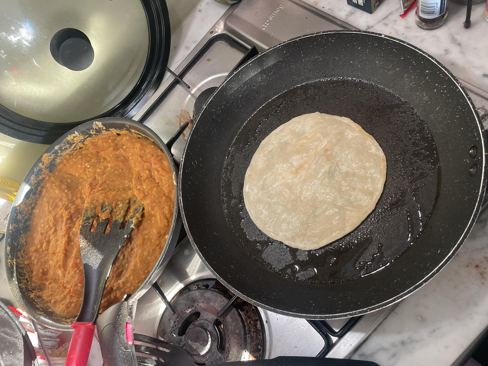 The image shows a close-up, overhead view of a kitchen scene, likely in a residential setting. A gas stovetop is visible, with a large black frying pan in the foreground containing a round, flatbread that is lightly browned and appears to be cooking in oil. To the left and slightly behind the frying pan, a larger pot or pan is seen, filled with a thick, orange-colored mixture that resembles mashed beans or a stew. A black and red spatula is resting in this pot, suggesting it is being used to stir or serve the contents. A portion of a silver lid with a black knob is visible on the right side of the frame. The stovetop itself shows signs of use, with some staining around the burners. The overall impression is of food being prepared for a meal. The image does not contain any specific visual cues that would allow for identification of a location within the city of Melbourne, Australia.