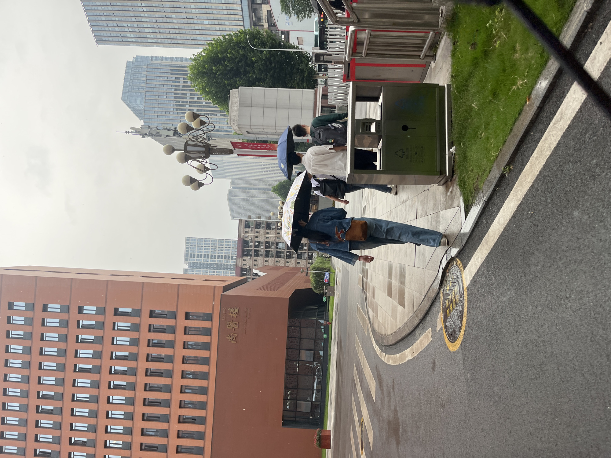 Four individuals are walking on a wet pedestrian sidewalk next to a street with a crosswalk. The person in the foreground on the left is walking away from the camera, holding a patterned umbrella. They are wearing a denim jacket and blue jeans. To their right, a person in a white shirt is holding a dark blue umbrella, and another person wearing a backpack and a dark green shirt is walking behind them. The fourth person, also carrying an umbrella, is partially visible behind the person in the white shirt. The environment features a tall, reddish-brown brick building on the left with many windows, and several modern, glass-fronted skyscrapers in the background. A street lamp with multiple globes is visible between the brick building and the skyscrapers. There is a green trash receptacle on the sidewalk. The ground is wet, suggesting it has recently rained. The scene appears to be in a city, possibly in Yuzhong District, China, given the architectural styles and the presence of Chinese characters on the building.