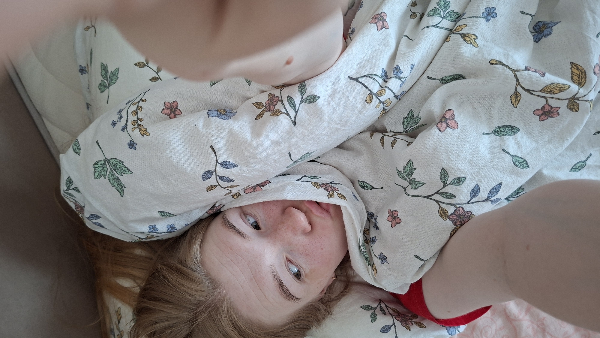 A woman with freckles and red hair lies in bed under a white floral-patterned comforter. Part of her face is visible, peeking out from the covers. Her eyes are open, looking to the side. The bed appears to be in a room with a beige wall. The woman's hand and arm are visible in the foreground of the shot. A red item is also visible near the woman's head, possibly a sock. The image does not provide enough information to determine the location in Chaikovsky, Russia, or any activities beyond lying in bed.