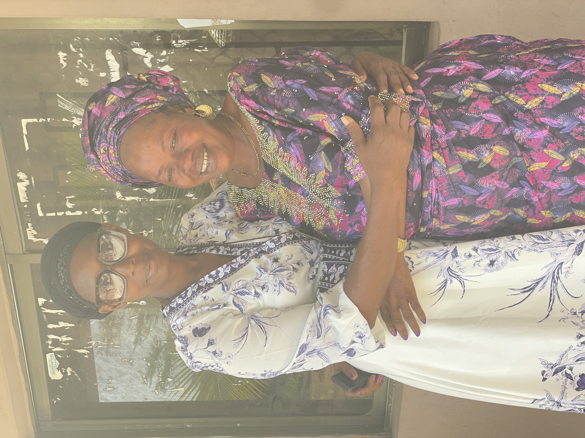 Two middle-aged women pose for a photo. The woman on the right, with a purple and black patterned head wrap and dress, is smiling broadly. The woman on the left, wearing glasses and a white dress with blue floral patterns, looks towards the camera with a slight smile. They are embracing in front of a reflective glass window that seems to look out onto a street in Badagry, Nigeria.