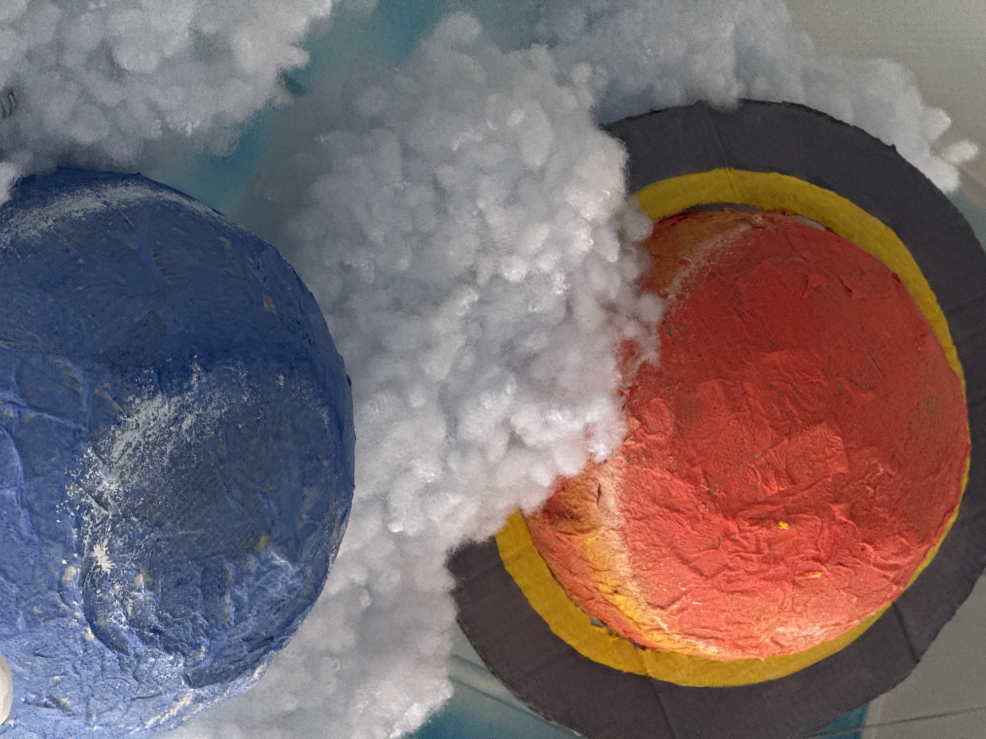 The image displays a close-up view of a handcrafted diorama or art piece featuring two spherical objects, likely representing celestial bodies, surrounded by fluffy white material.
On the left, a blue sphere with a textured, uneven surface is prominent. It has variations in tone and some white specks, suggesting a weathered or painted appearance, possibly depicting a planet like Earth with clouds or landmasses.
To the right, a larger, vibrant red sphere is depicted. This sphere also has a heavily textured, almost crumpled surface. It is encircled by two distinct, flat rings: an inner yellow ring and an outer dark gray or black ring. This design strongly suggests it represents the planet Saturn.
Large masses of white, fluffy material, resembling cotton batting or artificial snow, fill the space between and around the spheres, creating a cloudy, ethereal, or cosmic atmosphere. A lighter blue background is visible in the upper and lower areas of the composition, possibly serving as a sky or distant space. The objects appear to be made from craft materials such as papier-mâché or painted foam, giving them a tactile, handmade quality. The scene is static, depicting an artistic interpretation of planets. There is no visible text.