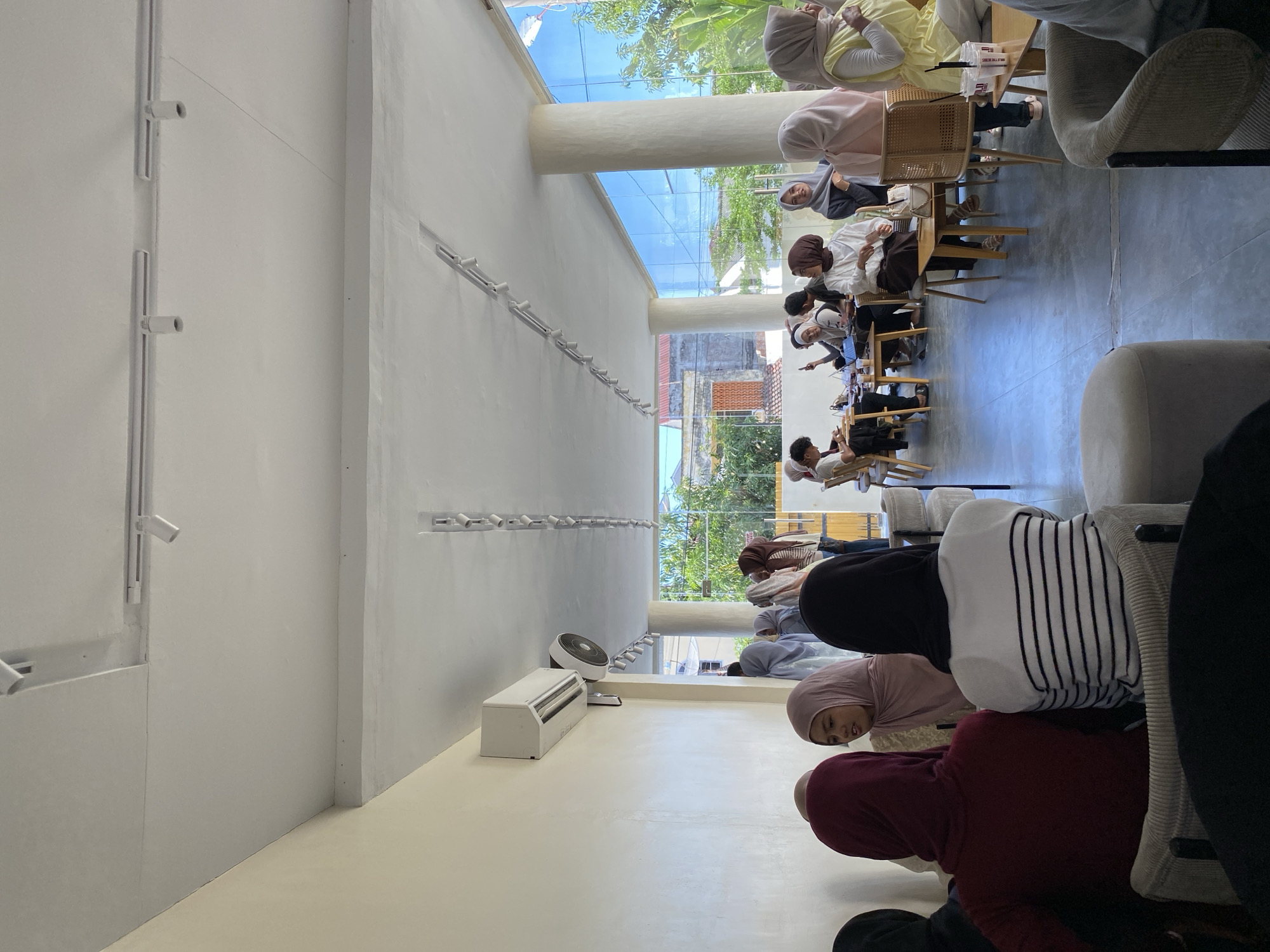 The image depicts a group of people, most of whom are wearing hijabs, gathered in what appears to be a cafe or meeting space in Lhokseumawe, Indonesia. The cafe is a modern space with white walls and ample natural light. Overhead, there is a row of track lighting installed along the wall. In the background, more people are seated at tables, engaging in conversation and consuming drinks. In the lower half of the image, a group of people appear to be seated on the floor. An air conditioning unit is placed on the wall near the floor. A large fan is on in the background behind the people on the floor. The environment appears modern and well-lit.