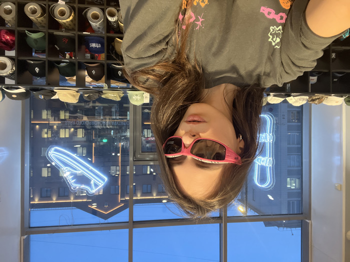 A young female, estimated to be in her 20s, is captured in a selfie within a retail establishment located in Krasnoyarsk, Russia. She has long, dark hair and wears pink, wrap-around sunglasses. Her attire consists of a dark grey long-sleeve t-shirt with various abstract, colorful graphic designs, including pink chain links and orange shapes. The subject is centered in the frame, looking directly towards the camera with a neutral expression.
The interior background reveals shelves displaying numerous baseball caps, some bearing recognizable brand logos such as Volcom and Etnies. Below these displays, rolls of material, possibly fabric or wrapping paper, are neatly stacked on lower shelves.
Through large windows behind the subject, an urban exterior scene is visible at what appears to be dusk or early night. The sky is dark blue. Two prominent neon signs, one shaped like a snowboard and another like a sneaker, both emitting a blue light, are suspended within the window frame. Across the street, multiple multi-story buildings with illuminated windows are visible. Partial Cyrillic text reading "УРАТУРА КРАСНОЯРСКОГО КРАЯ" is discernable on a structure or sign in the background, confirming the location in Krasnoyarsk, Russia.