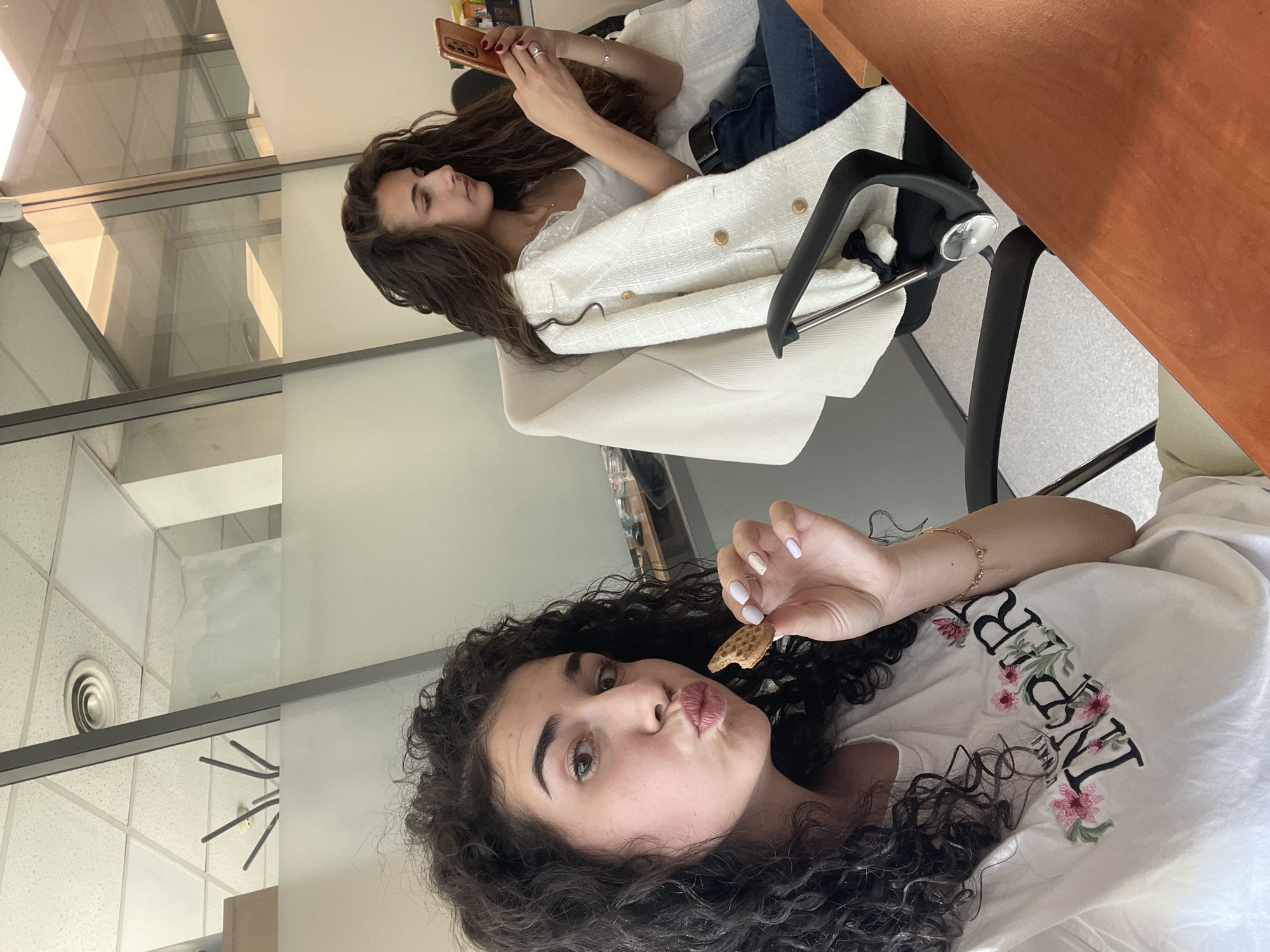 Two women are visible in the image. One woman with dark curly hair is in the foreground, holding a cookie, and posing with pursed lips. She wears a white t-shirt with floral embroidery on the left side of the chest and a gold bracelet. Behind her, another woman with long, wavy dark hair is sitting in a white chair and using a smartphone. She is wearing a white top, blue jeans, and a cream-colored tweed jacket is draped over the chair. The location appears to be an office space with visible windows and ceiling tiles. There is no definitive evidence indicating the specific location is within Sofia, Bulgaria. Further forensic examination may be needed to ascertain the exact location.