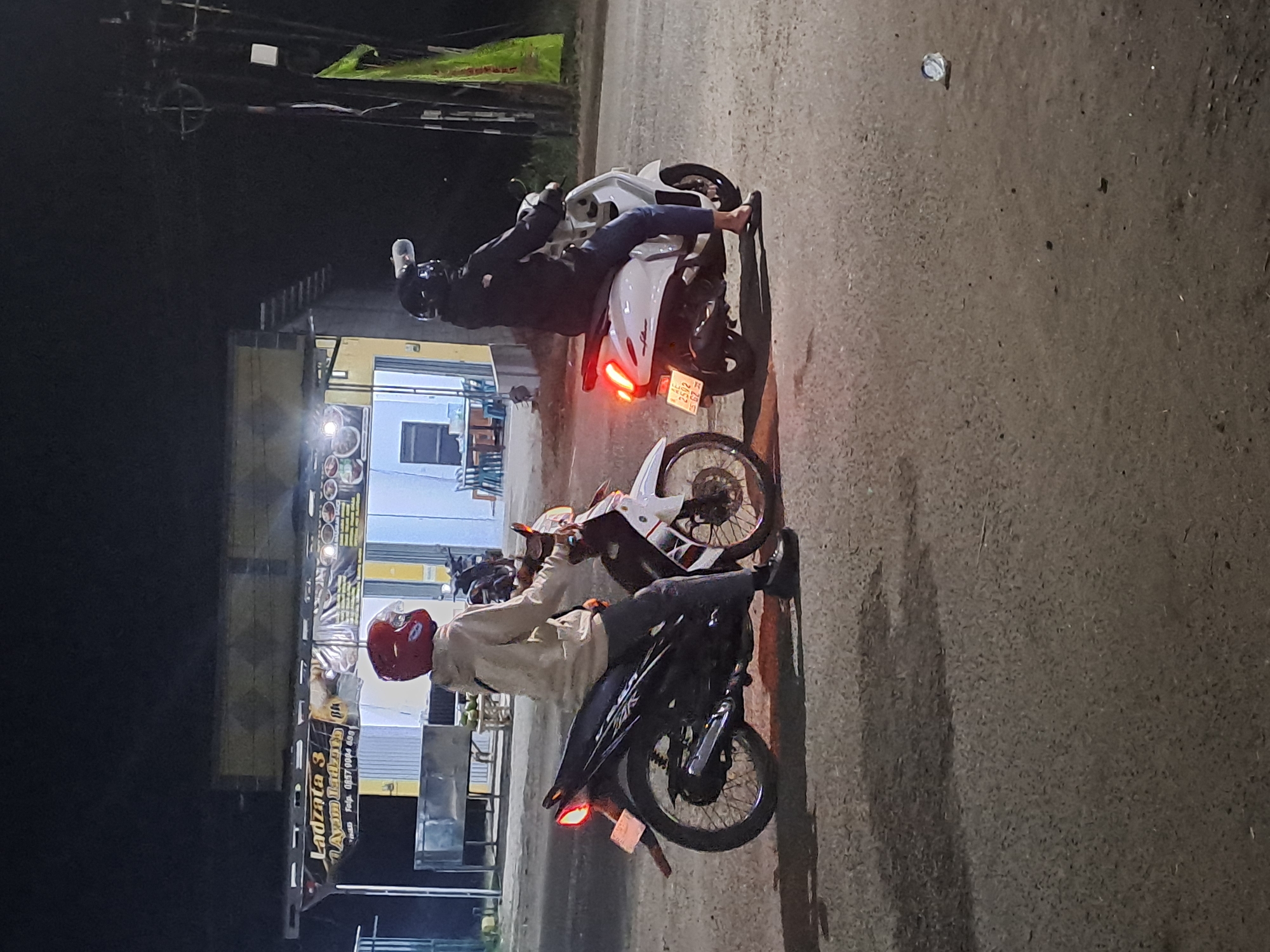 Two individuals on motorcycles are on a street at night, with a building in the background. The person on the left is wearing a red helmet and a light-colored hoodie, riding a dark-colored motorcycle. The person on the right is wearing a dark helmet and jacket, riding a white scooter. The building in the background appears to be a restaurant or food establishment, with a sign that reads "Ladzata 3 Ayam Ladza" and a phone number. The scene is dimly lit, suggesting it is nighttime. The environment appears to be urban or semi-urban, with a paved road and some overhead utility lines visible. The overall impression is a casual scene of people on motorcycles at night in what appears to be a commercial area in Slambur, Indonesia.