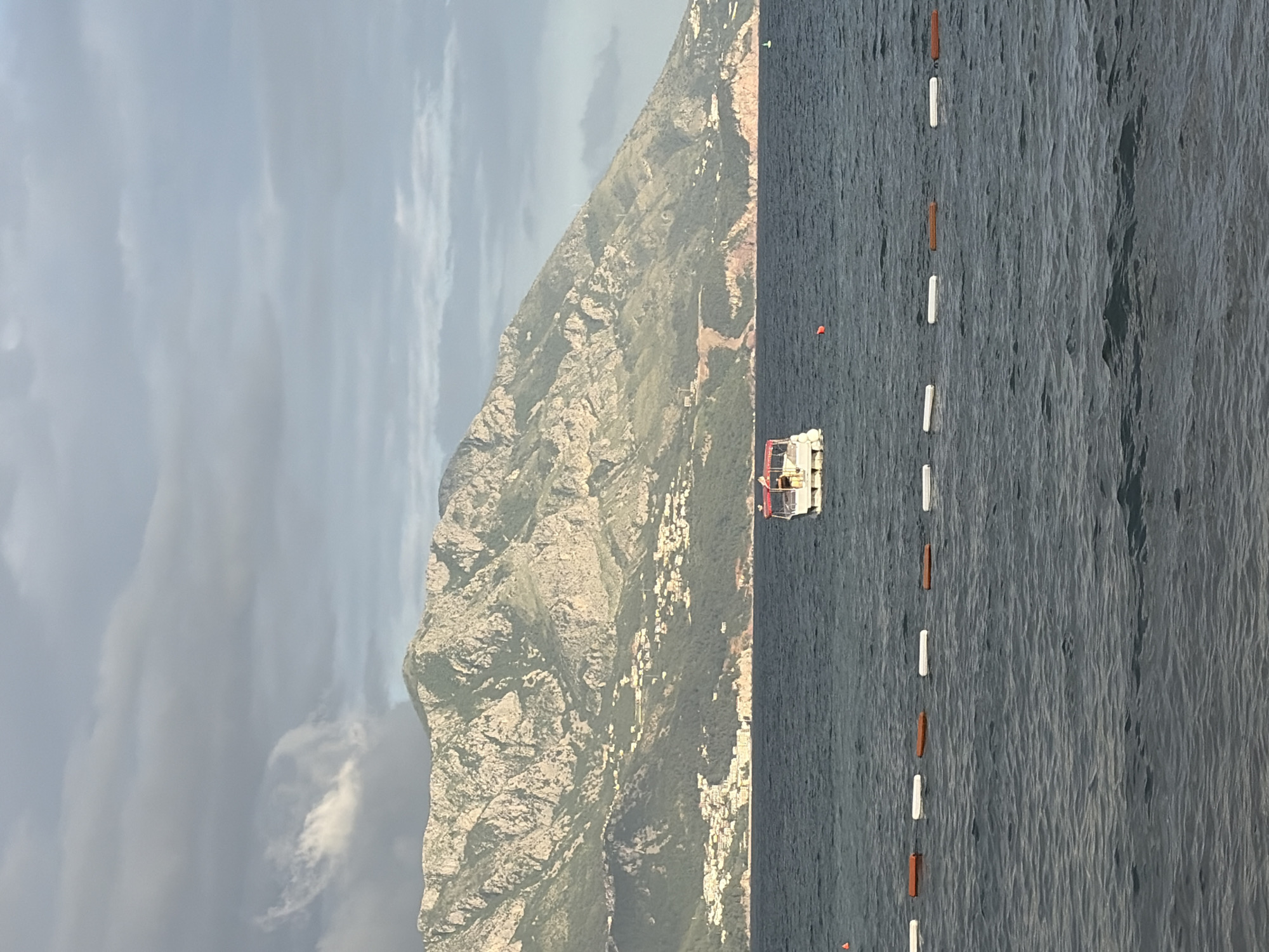 The image depicts a coastal scene, likely near Budva, Montenegro. On the left, a large, rocky mountain dominates the view, its slopes sparsely covered with vegetation and dotted with buildings. A cloudy sky looms overhead. To the right, the dark, rippled surface of the sea stretches out. A line of alternating white and orange buoys extends across the water, possibly marking a swimming area or boundary. A small white boat is visible in the water between the mountain and the buoys.