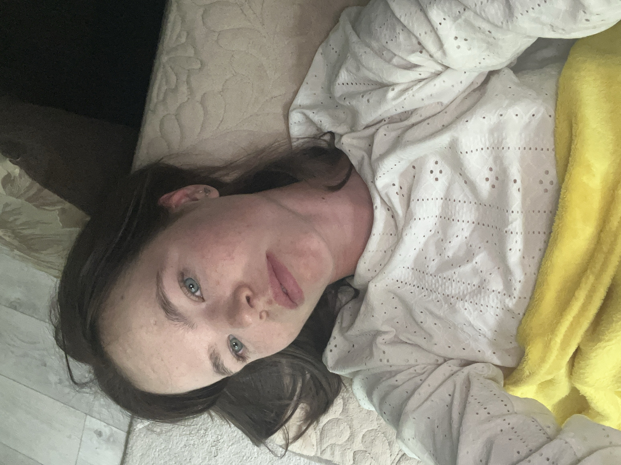 A person is lying on a bed. The person has light skin, blue eyes, and dark brown hair. They are wearing a white, lacy top and are partially covered by a yellow blanket. The bed has a beige, textured mattress. Part of a dark-colored object, possibly furniture, is visible to the left of the bed, and a portion of a light-colored floor is visible in the lower left corner. The setting is likely indoors, given the presence of the bed and other furniture. I am not sure if this location is in the city of Chaikovsky, Russia.