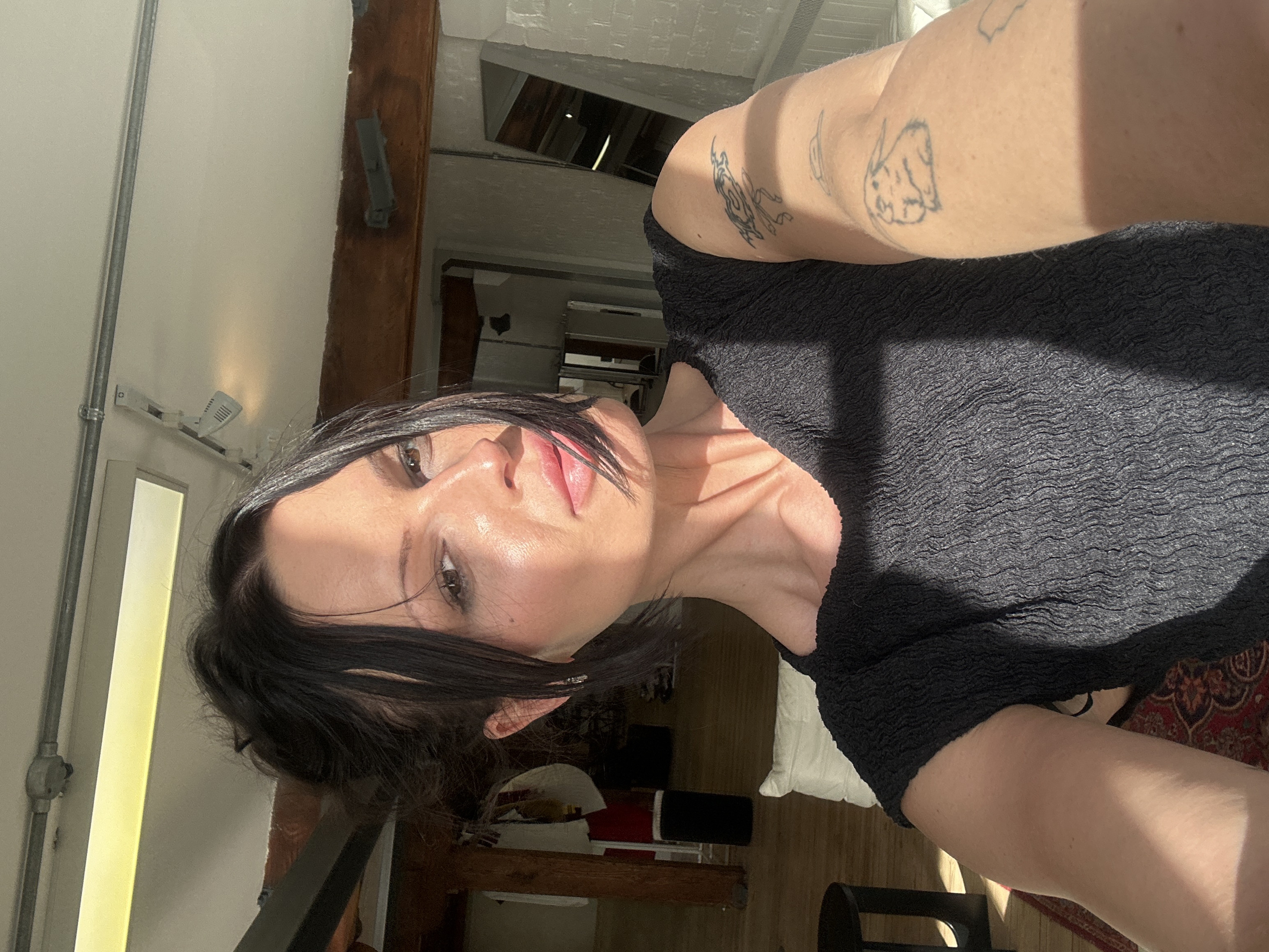 A single adult female is depicted in a close-up selfie within an indoor setting in the City of London, United Kingdom. The subject, with dark hair parted and partially sweeping across her face, has brown eyes and pink-toned lips, exhibiting a neutral expression with a slight pout. She wears a dark, short-sleeved top with a crinkled texture. Direct sunlight illuminates the right side of her face, neck, and right arm, creating distinct shadows. Her right arm displays three linear tattoos: an abstract design on the upper arm, a small, simple shape below it, and a rabbit figure on the forearm. The background features exposed wooden ceiling beams and a white ceiling with visible metal conduit and fluorescent light fixtures. White brickwork is visible on the right wall, where a full-length mirror leans. A dark, circular stool, a white cushion, and a patterned red and black rug are present on the floor to the left. Additional obscured objects, including hanging fabrics, are visible in the deeper background.