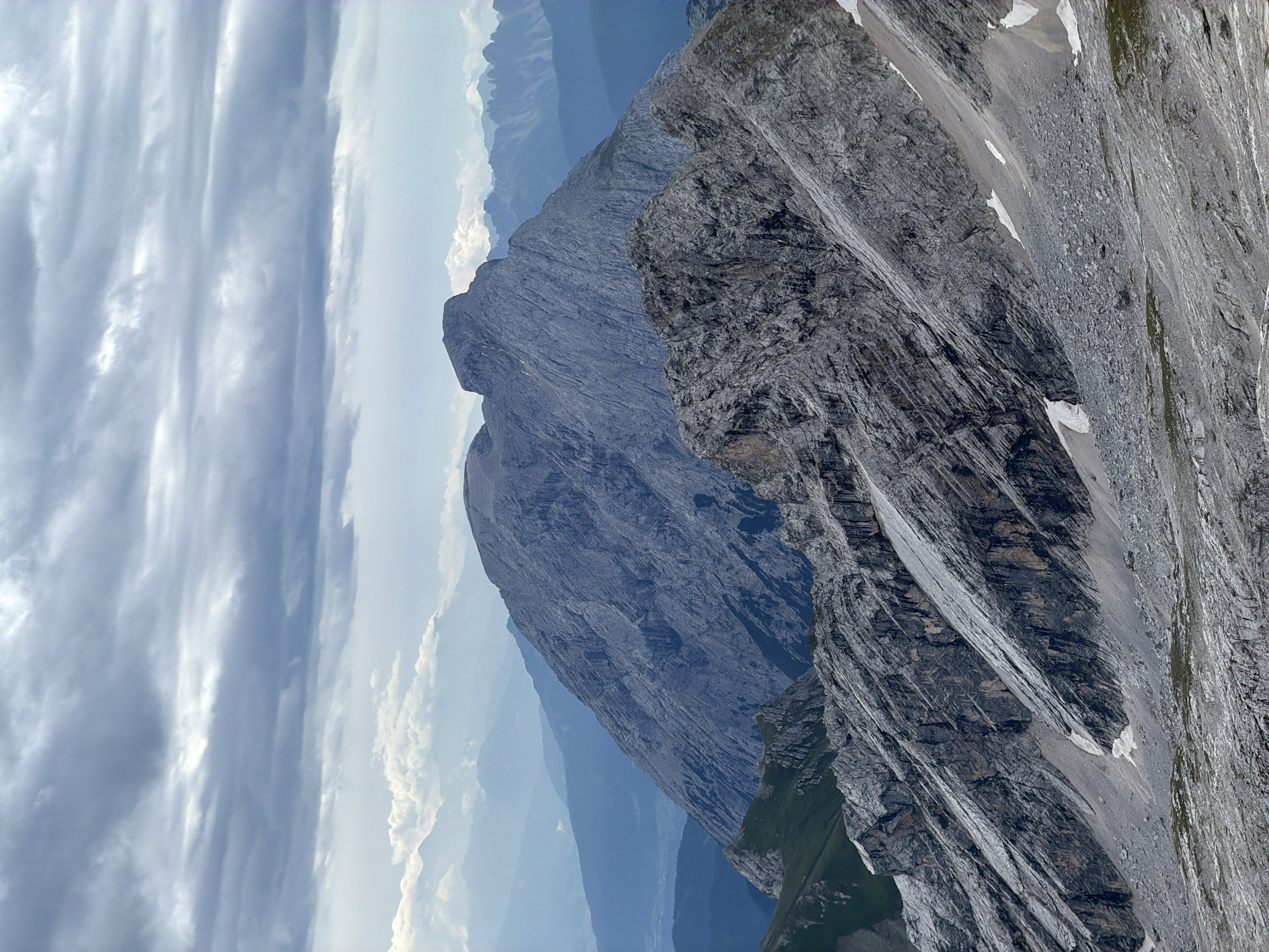 This image depicts a panoramic view of a rugged mountain landscape under a cloudy sky. The foreground is dominated by a steep, rocky slope with visible striations and scree. In the midground, another, larger mountain peak rises, its gray, textured surface showing the effects of erosion. In the background, multiple layers of mountains recede into the distance, their colors becoming progressively bluer and hazier, suggesting depth and atmospheric perspective. The sky is filled with heavy, gray clouds, with breaks allowing for glimpses of lighter sky and distant mountain ranges. There are no people or man-made objects visible in the scene. The image is a high-angle shot, likely taken from a higher vantage point, looking out over the mountainous terrain. The scene is natural and undeveloped.