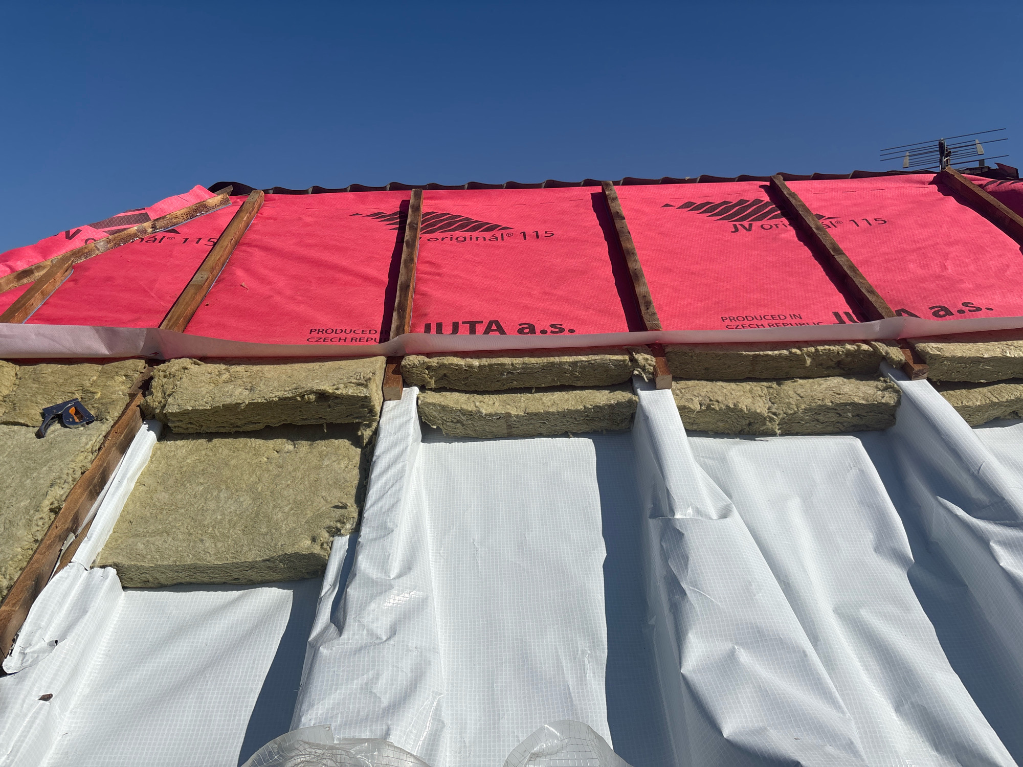 The image depicts the construction of a roof in Zhavoronki, Russia. The scene focuses on the exposed roof structure, showing wooden rafters supporting a layer of pink waterproof membrane with "JV original 115" and "JUTA a.s." branding. Beneath this membrane, sections of yellowish insulation material are visible, secured by wooden battens. A white, grid-patterned waterproof sheeting is being installed below the insulation. The sky above is clear blue. There are no people visible in the frame.