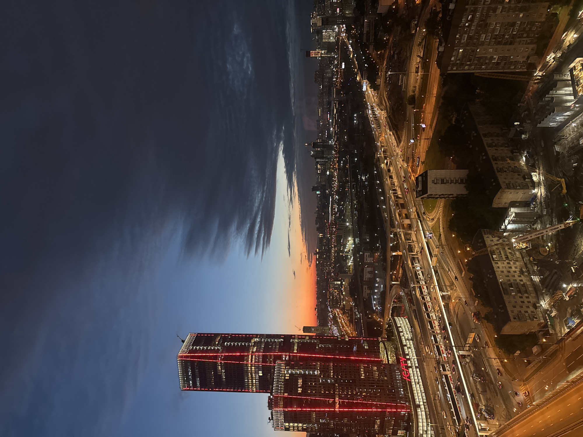 An aerial view captures a city skyline at dusk in Khimki, Russia. Dominating the left of the frame is a brightly lit skyscraper with red vertical accents, flanked by a shorter, similarly illuminated building. Below them, a complex network of multi-level highways is visible, with headlights and taillights indicating ongoing traffic. Several residential apartment buildings are interspersed throughout the landscape. In the background, a vast urban expanse stretches towards the horizon, dotted with illuminated windows and streetlights, under a sky transitioning from a vibrant orange sunset near the horizon to a deep blue and dark gray cloud cover overhead. Construction sites with cranes and scaffolding are present in the foreground and mid-ground. The overall scene depicts a bustling urban environment during the twilight hours.