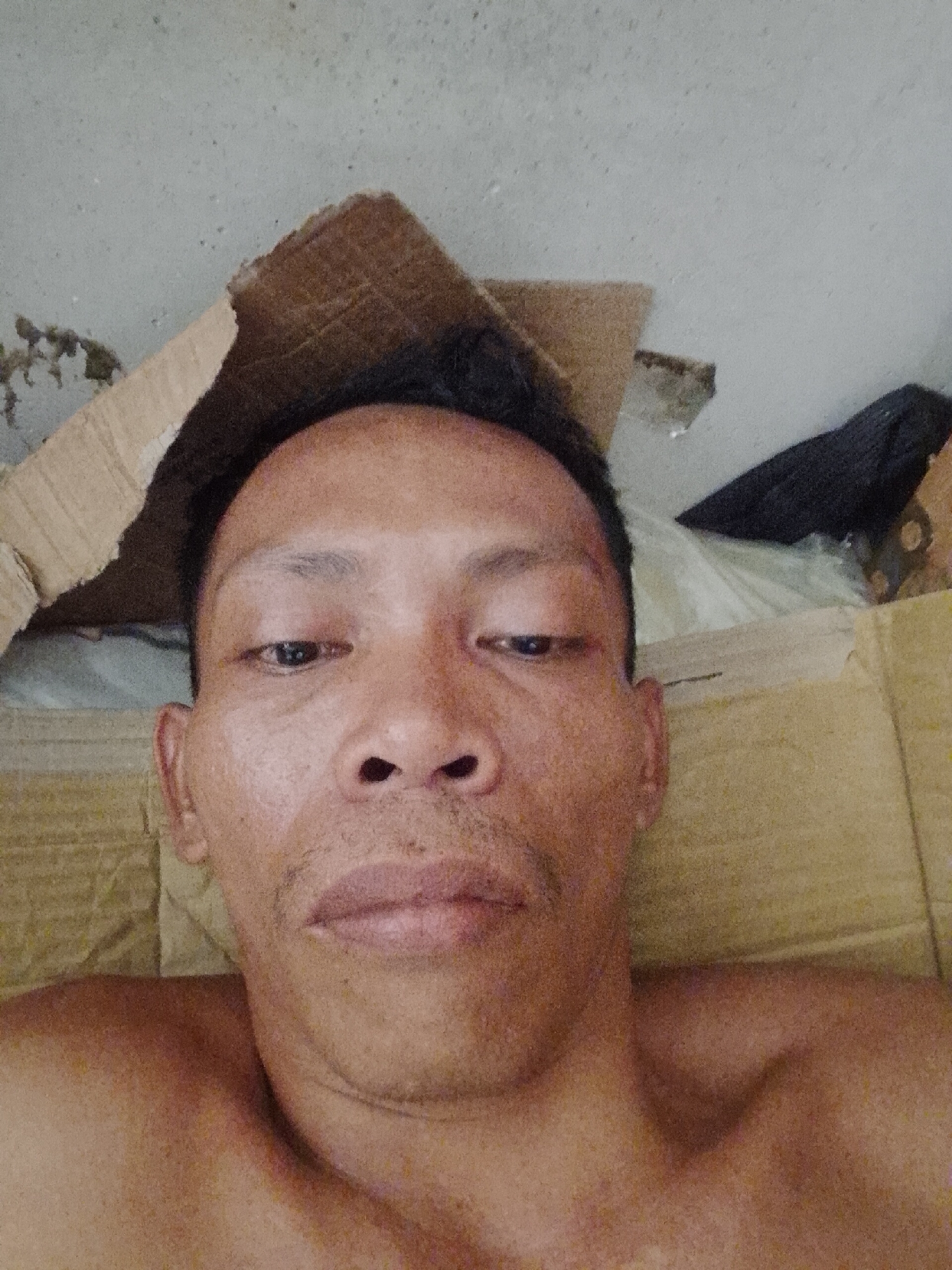 A close-up, high-angle self-portrait of a bare-chested middle-aged Asian man with short, dark hair. He has brown eyes, a wide nose, and thin mustache hairs. His skin is slightly weathered with visible pores. The background consists of a concrete surface and discarded cardboard, giving the impression that he is lying on the floor or a makeshift bed. There's also a glimpse of a dark cloth or fabric object. The image was presumably taken indoors in Bukit Ketok, Indonesia. No actions or interactions can be observed.