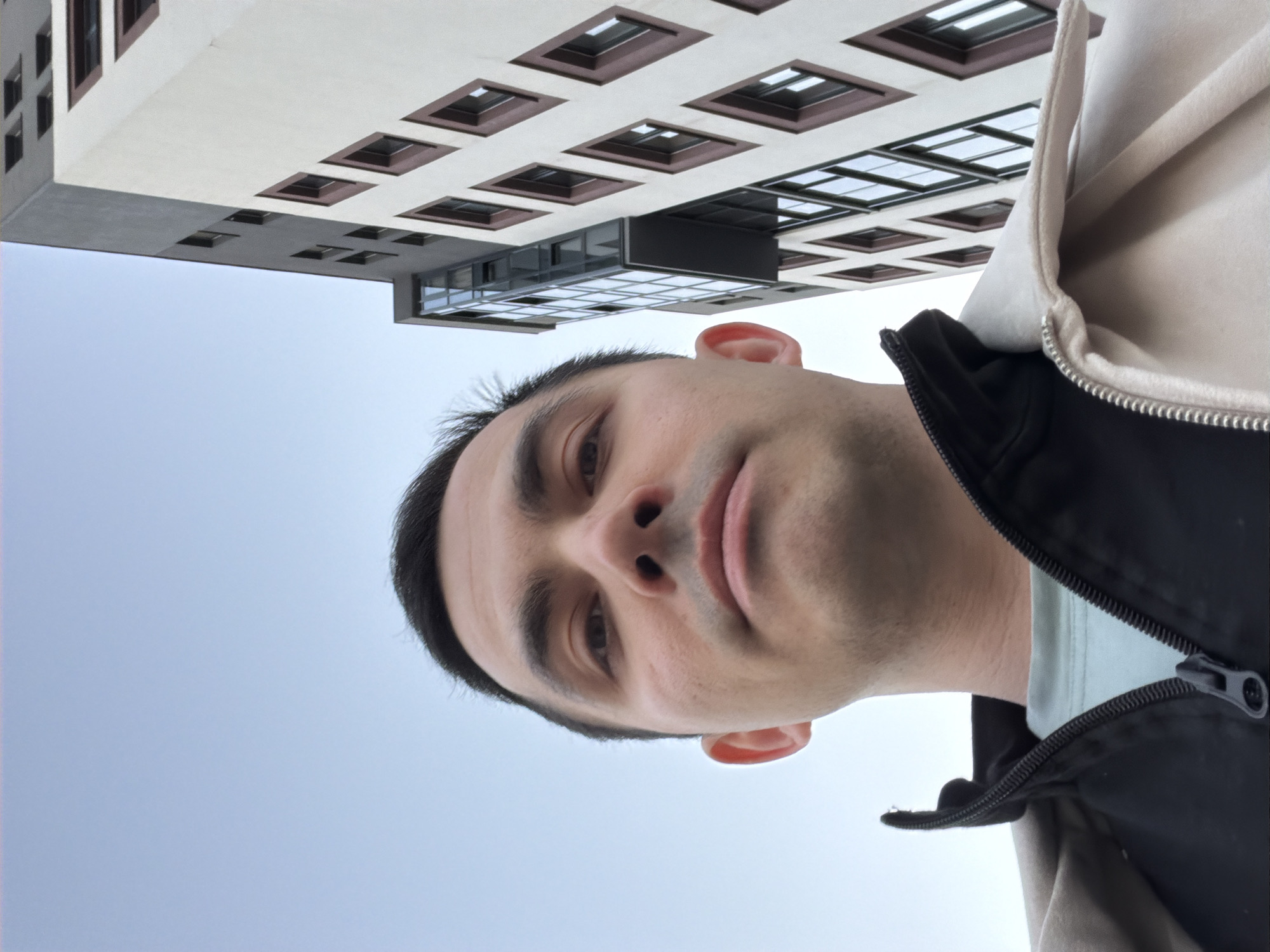 A man is looking at the camera. He is wearing a jacket. Behind the man is a building with many windows. The background is the sky.