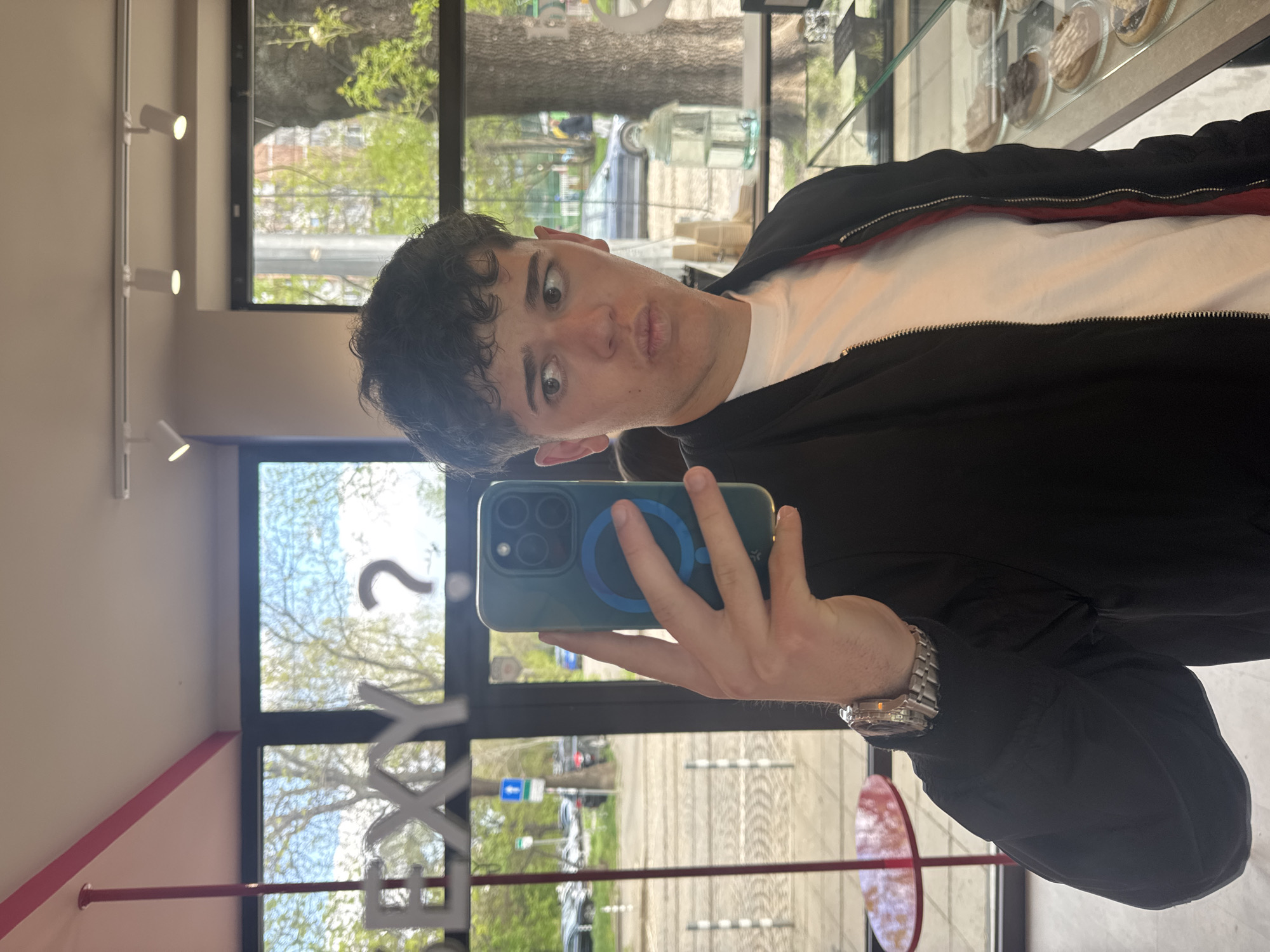 A young man takes a selfie in what appears to be a cafe or shop, captured from a slightly low angle. He has dark, curly hair and is looking directly at the camera with a playful expression, featuring wide eyes and puckered lips. He is wearing a white T-shirt under a black jacket with a red stripe detail on the collar. On his left wrist, he wears a silver watch with a dark face. He holds a teal-colored smartphone with a visible blue MagSafe ring on its back, obscuring part of his face.
The setting is indoors, with natural light streaming through large windows that line the background. Outside, green trees suggest spring or early summer, under a clear blue sky. A street with cars and signs is visible through the window. Inside, a light-colored wall with white track lighting fixtures runs along the left. To the man's right, a display case containing baked goods or desserts can be seen. A red, circular table is partially visible in the bottom right, reflected in the window. A distinct pink accent line runs along a wall corner.
Visible text on the window reads "EXY" and a partial question mark shape. The scene is brightly lit, indicating daytime. The man's expression conveys a lighthearted and humorous mood. The presence of a cafe setting and street view aligns with the given context of Sofia, Bulgaria.