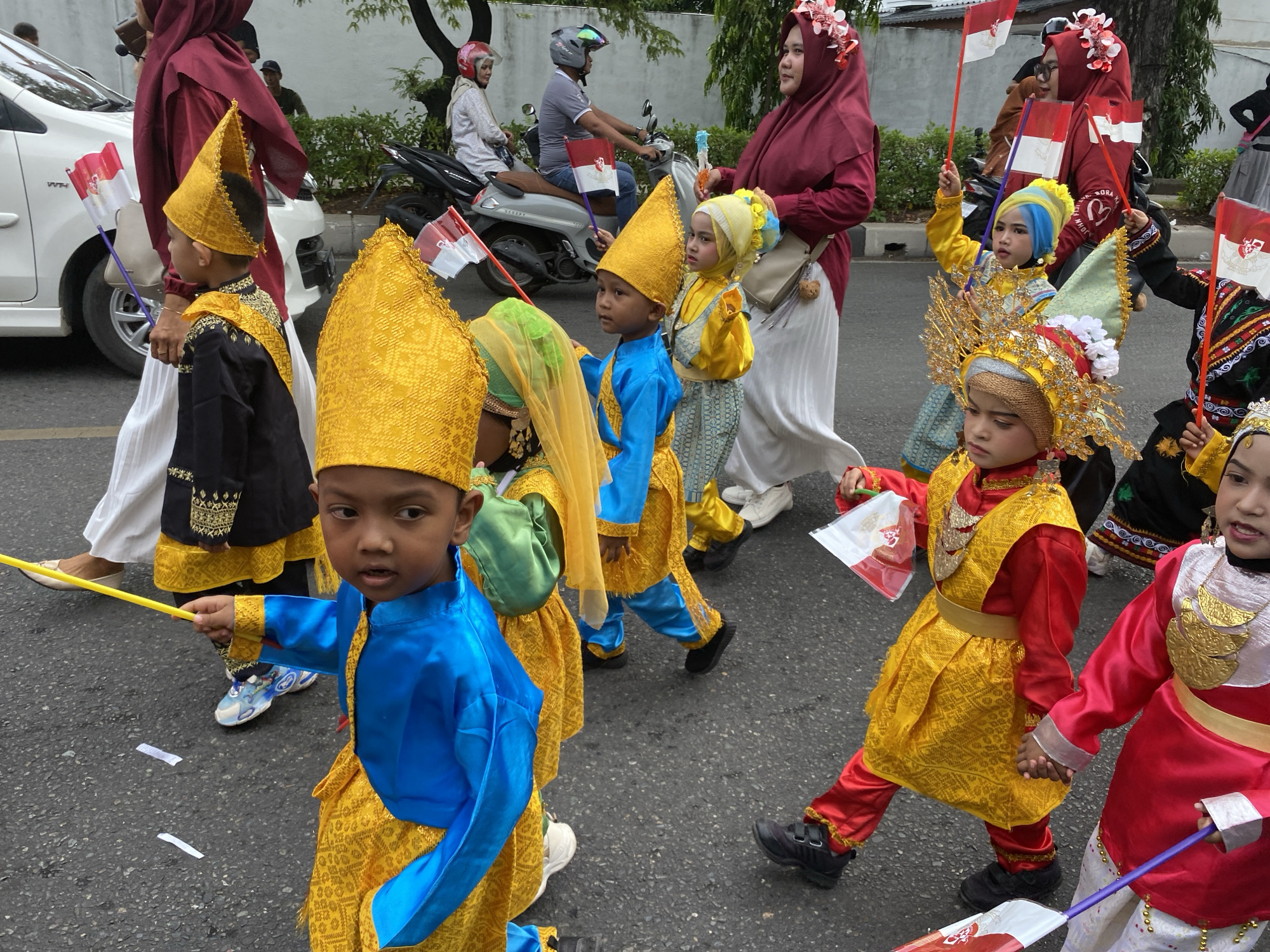 In Banda Aceh, Indonesia, a group of children in traditional attire, some wearing golden headwear and others in vibrant blue and yellow outfits, participate in a parade. They are walking on an asphalt road, and some are holding small Indonesian flags. Adults in maroon headscarves and white skirts are visible in the background, accompanying the children. A white car and a motorcycle are parked nearby, and trees line the street. The children's clothing features intricate patterns and gold embellishments. The atmosphere appears festive, suggesting a cultural celebration or event.