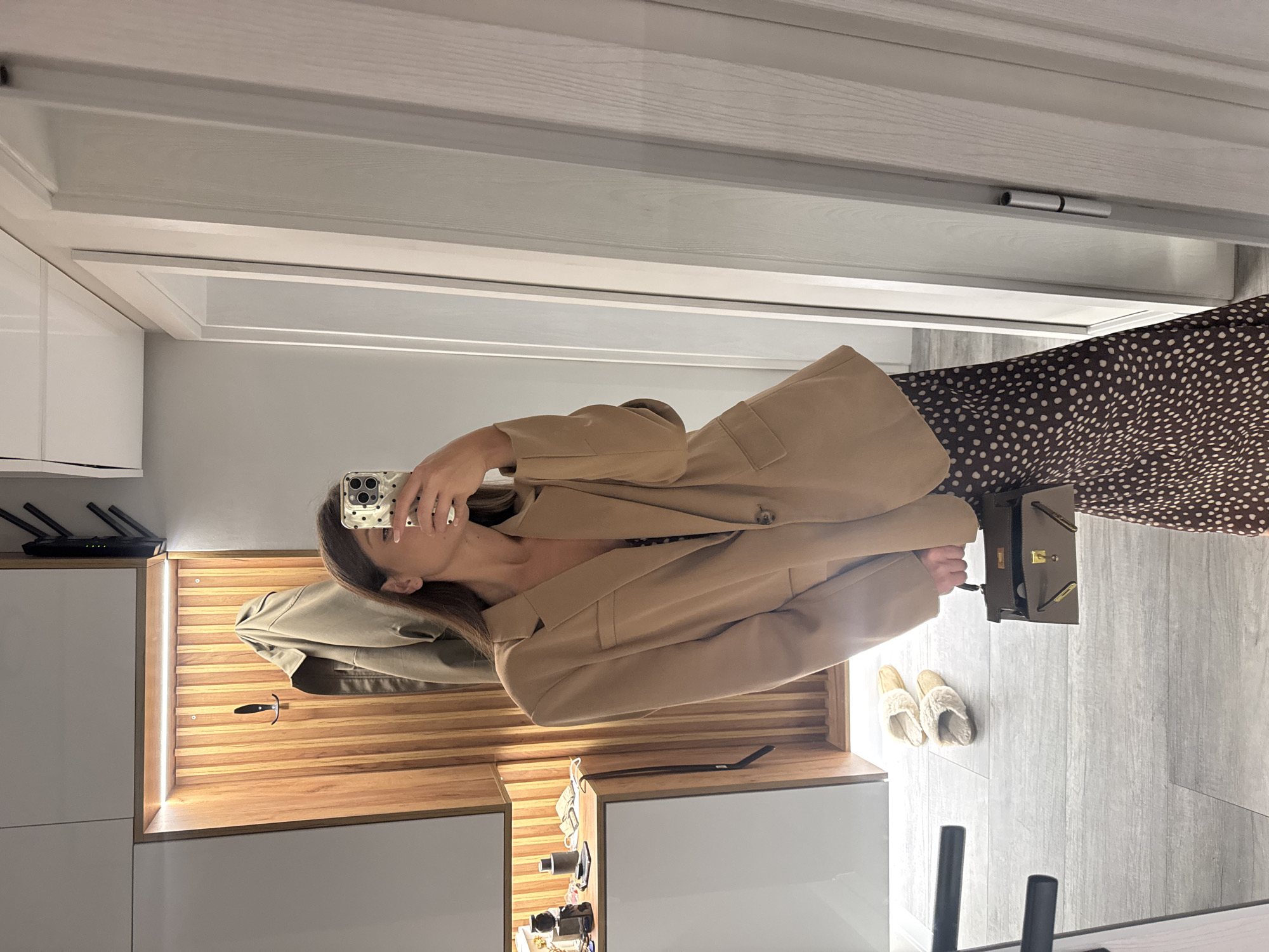 A woman in a tan blazer and brown polka dot dress is taking a selfie in what appears to be an indoor setting, likely her home. She is holding her phone in her right hand. A light-colored wooden cabinet with what appears to be interior lighting is visible. Next to the cabinet, there are household items such as a black router and other personal care products and decorative items. A pair of slippers and a dark handbag are near the woman on the floor. The floor is a light gray wood-patterned tile.
There is no information available in the image to determine a specific location in Burgas, Bulgaria.