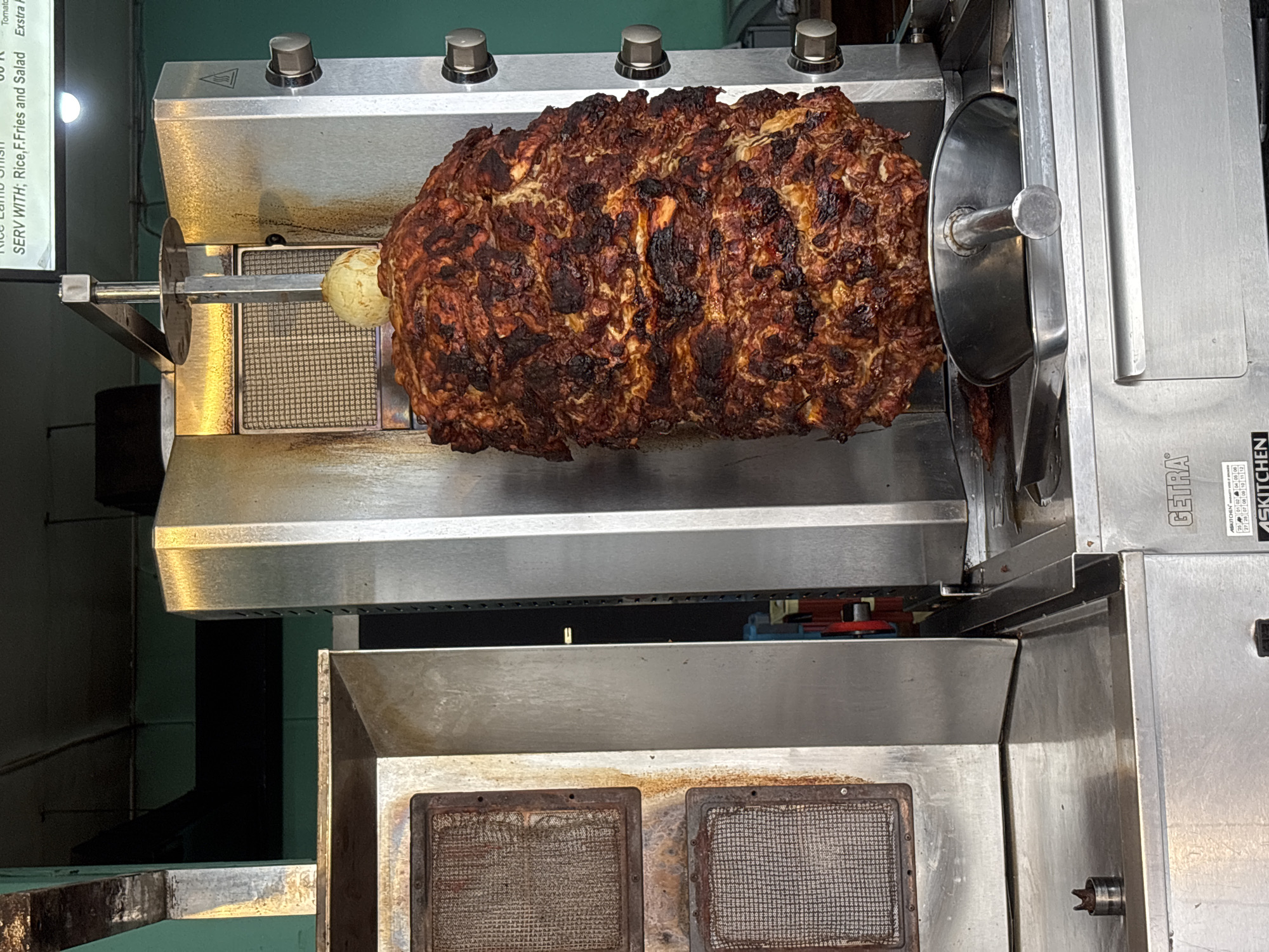 A large mass of cooked meat, likely shawarma or gyro meat, is mounted on a vertical rotisserie grill. The meat is a dark brown color with some charred areas, suggesting it has been cooking for some time. The grill itself appears to be stainless steel and features metal mesh screens positioned below the meat. There are several control knobs on top of the grill. A sign nearby mentions the dish can be served with rice, fries, and salad and is found in Kuta Selatan, Indonesia.
