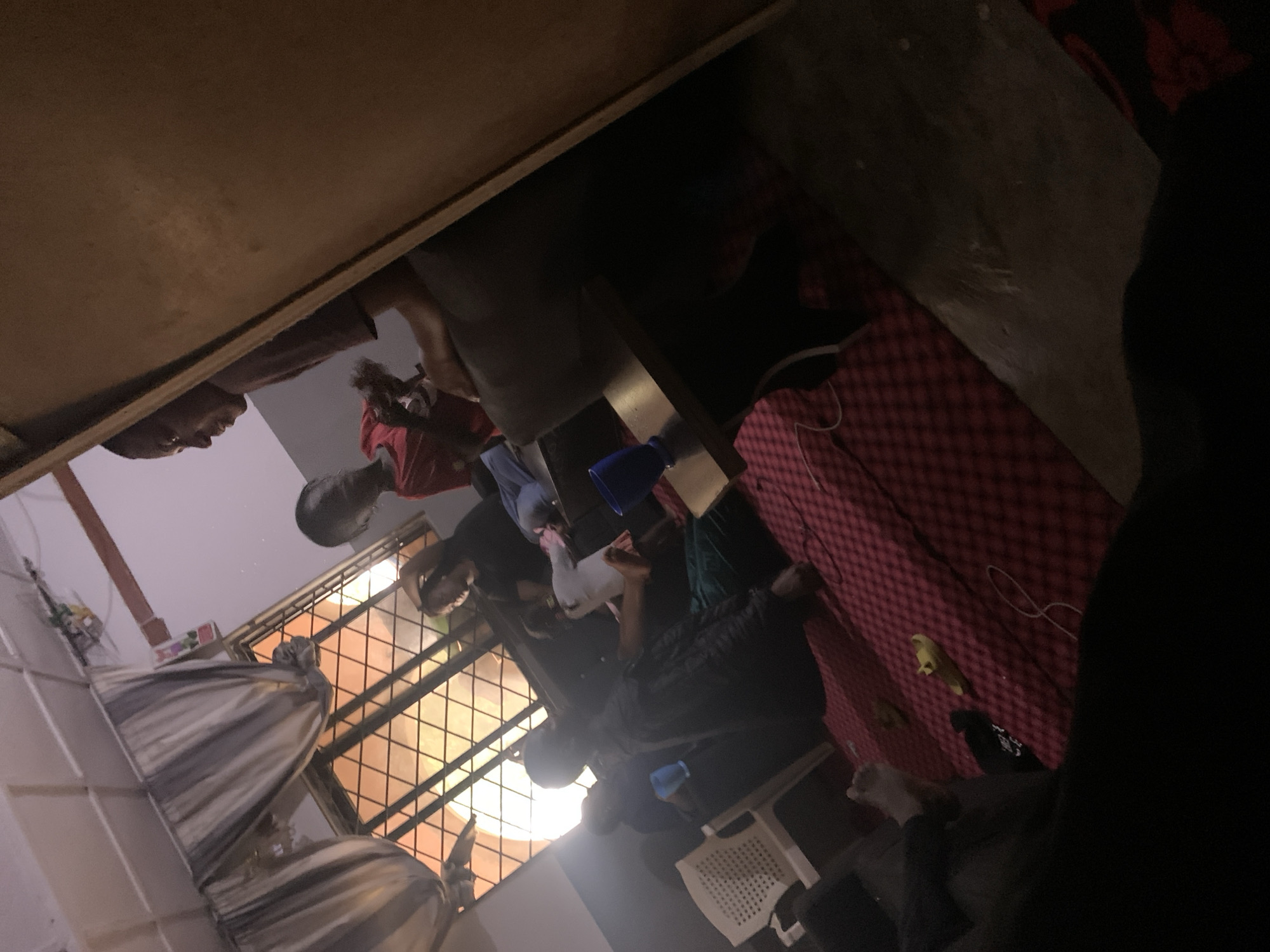 This media file depicts a group of people in an indoor setting, likely a room or common area, in Jos, Nigeria. The image is taken from a low, wide-angle perspective, looking upwards. Several individuals are visible, appearing to be young men and women, in various casual poses. Some are seated, while others are standing or leaning.
The scene is dimly lit, suggesting it might be evening or night. The background includes elements like a window with a lattice or security bars, possibly with orange light filtering through, and a bed or mattress with a red checkered pattern. There are also some fabric items, possibly bedding or curtains, with striped patterns.
The activity appears to be informal social gathering. Some individuals are looking at their phones, while others are interacting or simply present in the space. One person in the foreground, wearing a red shirt, seems to be holding a phone and possibly taking a picture or video. Another individual, wearing a black shirt, has their arm raised. The general atmosphere seems relaxed.
Notable details include the rough texture of the ceiling or upper wall, which appears to be made of wood or a similar material. The presence of a white plastic chair and some blue objects, possibly containers or cups, adds to the casual setting. There is no visible text in the image. The scene appears to be a typical domestic or communal living space in Nigeria.