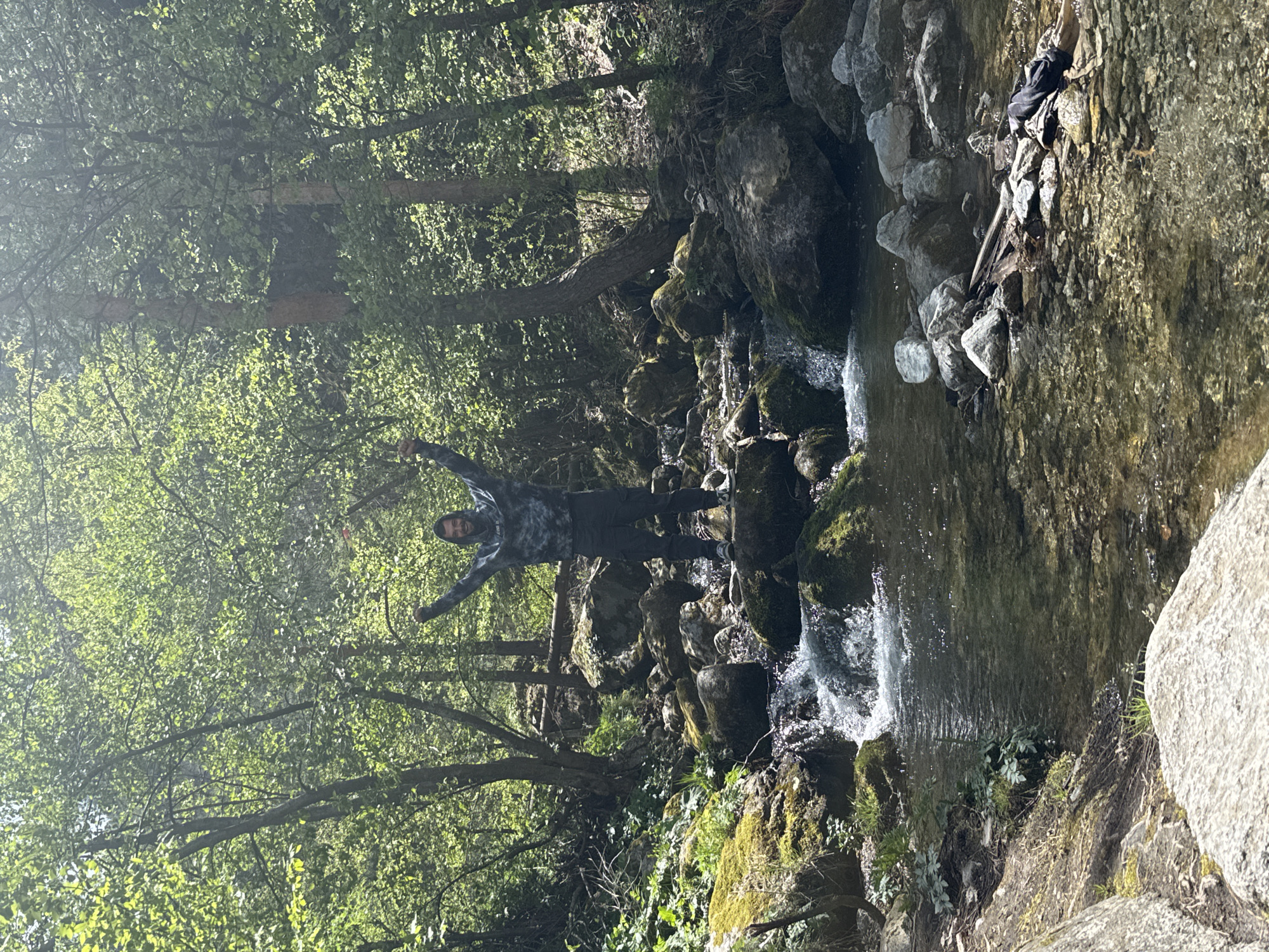 A person stands on mossy rocks in a creek bed in a densely forested area, with their arms outstretched. The location is Ovchartsi, Bulgaria. The creek cascades down rocks forming small waterfalls. The person is wearing a dark hooded sweatshirt and pants. There are trees with green foliage on either side of the creek. A large boulder is on the right side of the image. A backpack lies on the rocks next to the creek.