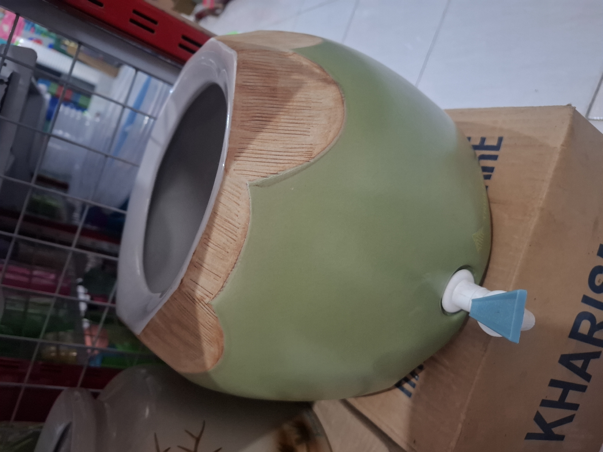A close-up, angled shot shows a coconut-shaped ceramic dispenser with a light green body and a tan, textured top. A white spigot with a blue handle protrudes from the lower front of the dispenser. The dispenser rests on a cardboard box with dark blue text partially visible. In the background, a light-colored vase or pot with a decorative pattern is to the left, and a metal shelving unit with various items behind a wire grid is to the right. The scene appears to be indoors, possibly in a store or warehouse, in Sumberwudi, Indonesia.