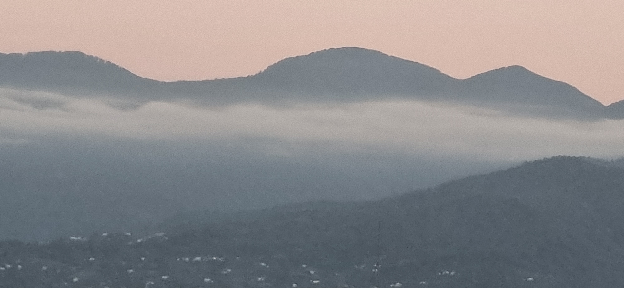 The image is a wide shot of mountainous terrain at dusk. The sky is a pale pink, transitioning to a light gray horizon. Several layers of dark blue-gray mountains are visible, with the furthest range appearing as a hazy silhouette against the sky. A thick band of white fog or low-lying clouds stretches horizontally across the middle ground, obscuring the lower portions of the mountains. In the foreground, the lower slopes of the mountains are densely covered with dark green trees. Scattered across the bottom of the image are small, white structures, likely houses, indicating a settlement. A single, thin, dark vertical line, possibly a tower or a pole, is visible rising from the tree line in the foreground. The overall impression is one of a peaceful, natural landscape as day turns to night. This scene is consistent with views from higher elevations overlooking the outskirts of Batumi, Georgia, a city known for its coastal location backed by steep, forested mountains.