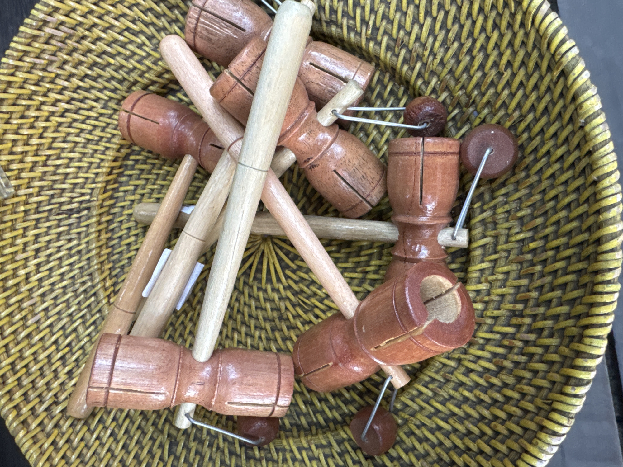 This image shows a collection of wooden toys, likely a variation of a click-clack or diabolo toy, nestled within a woven basket. The toys are predominantly made of polished brown wood, with some featuring lighter wood sticks. Each toy consists of a cylindrical or barrel-shaped wooden piece that has been carved to create a hollow interior and a series of slots. Thin metal wires extend from these wooden pieces, each terminating in a small, dark brown wooden bead or sphere. The basket is made of tightly woven natural fibers, predominantly yellow and green in color, creating a textured background for the toys. The lighting suggests an indoor setting, possibly a market stall or a home in Kuta, Indonesia, where such handcrafted items are commonly sold. There are no people visible in the image. The arrangement of the toys suggests they are being displayed or stored.