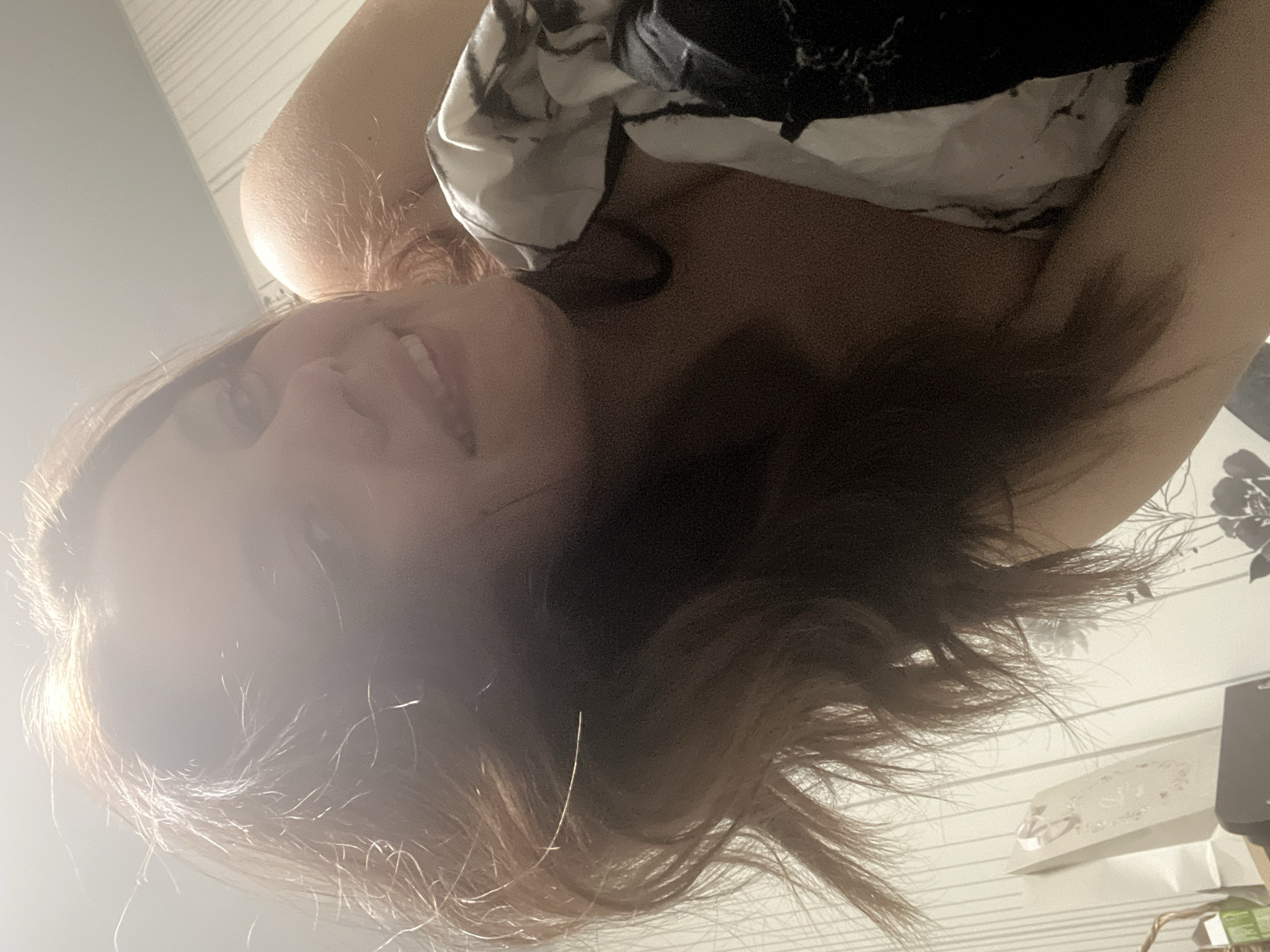This image, provided in its original rotated orientation, captures a low-angle selfie of a young woman indoors. When viewed correctly (rotated 90 degrees clockwise), the woman's head is on the left, and her body extends to the right.
The central subject is a young woman with reddish-brown, medium-length hair. She is smiling broadly, showing her teeth, and looking towards the camera. Bright light streams in from the left side of the frame, illuminating her face and hair and causing some overexposure in that area. She appears to be wearing a dark-colored top with subtle light-colored patterns or designs. Her left arm is visible, bent at the elbow.
The background indicates an indoor setting, likely a room in Chaikovsky, Russia. A patterned wall or wallpaper is visible, featuring horizontal lines and some floral or leaf motifs on the right side. On the right, there are hints of furniture or objects, including a white box or package with very faint, unreadable text or a logo, and a dark object, possibly an electronic device or speaker. The strong, bright light suggests it is daytime. The woman's smiling expression conveys a sense of happiness or contentment.