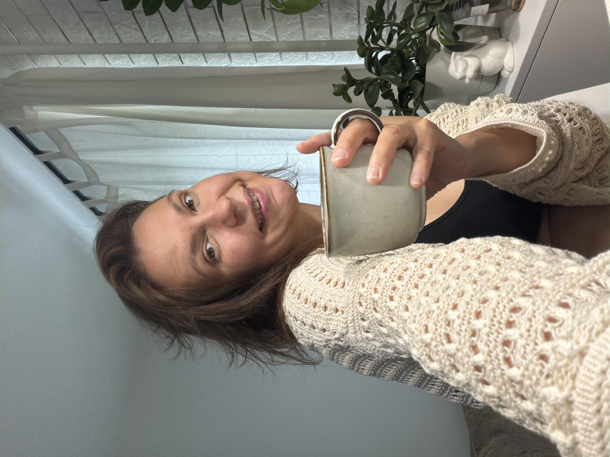 A woman with shoulder-length brown hair is holding a gray mug with a silver handle up to the camera. She is wearing a cream-colored, open-knit sweater over a black top. The background includes a window with white blinds, a green plant, and a white ceramic figure. The photo was taken indoors. The location is not discernible from the image. There is no evidence of Chaikovsky, Russia.