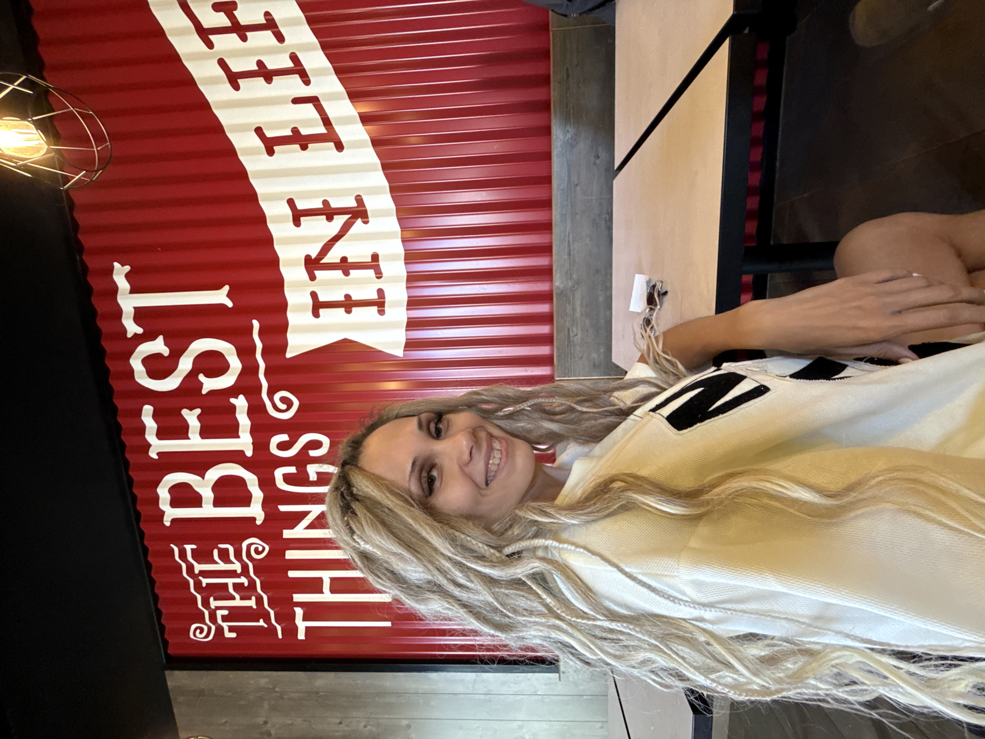 An indoor scene in Rusanovo, Russia, features a woman seated at a table, turned slightly to her right, facing the viewer with a smile that reveals dental braces on her upper teeth. She has long, wavy blonde hair with integrated braids, prominent eyelashes, and visible eye makeup. She wears an off-white long-sleeved top with a partial black, geometric design on the right shoulder. Her left hand rests on her left thigh. The background consists of a vibrant red wall with vertical corrugated panels. White text painted on this wall reads "THE BEST THINGS". A white banner design across the red panels displays additional text, partially visible as "IN LIF", suggesting "IN LIFE". Below the wall, a wooden bench or backrest is visible, with two light-colored tables with dark edges in the foreground. A pair of dark sunglasses and a small white rectangular object are present on the table closest to the woman. In the upper right of the frame, an industrial-style light fixture with an exposed bulb and a black wire cage hangs from a dark ceiling. A grey vertical structural element is visible on the far left.