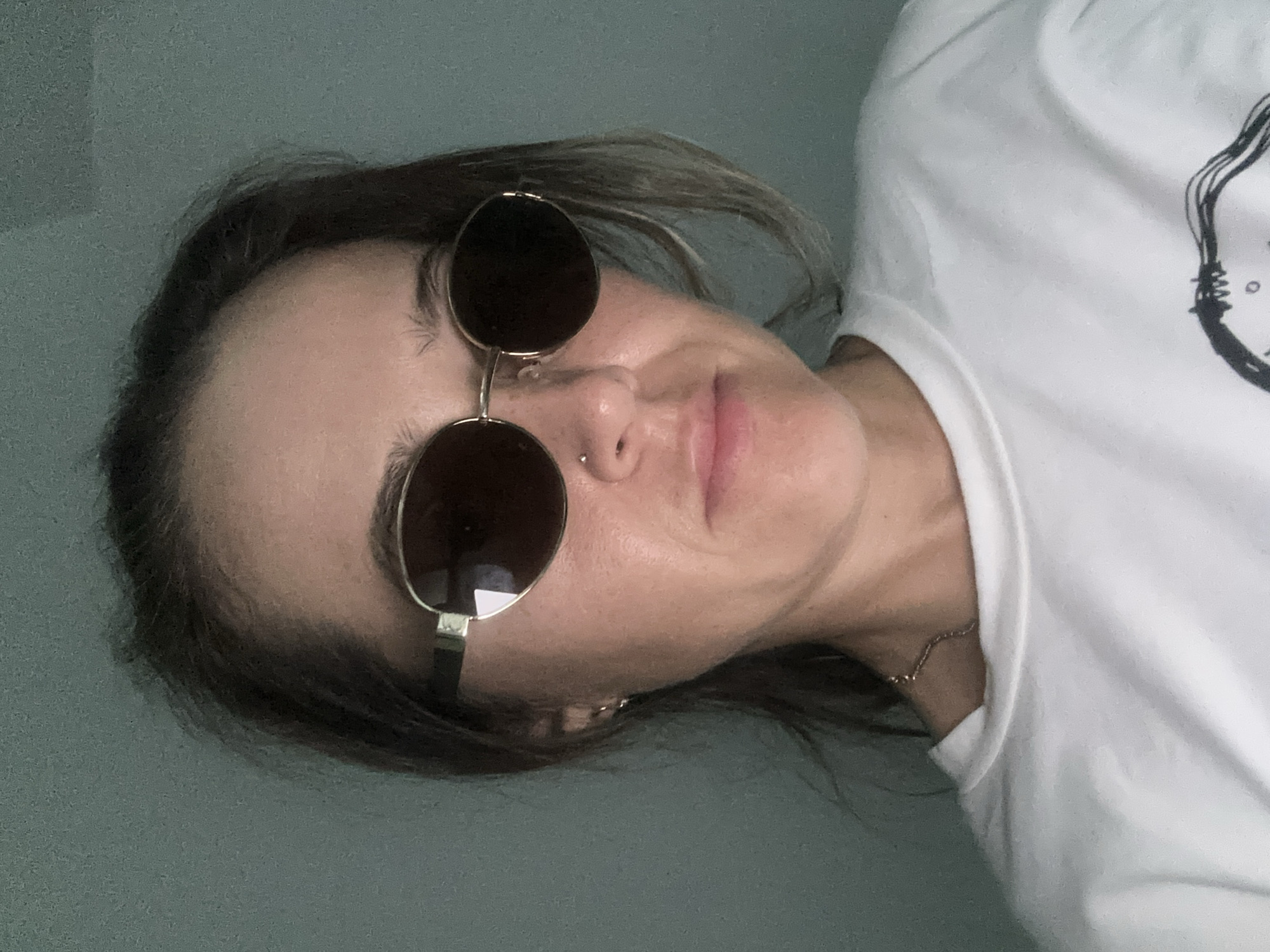 A woman with short, dark hair is wearing sunglasses and a white t-shirt with a design. She has a nose piercing and is wearing a delicate gold necklace. The background is a plain, muted green wall. The subject is likely posing for a selfie. There is no indication of the location being Bac Nha Trang Ward, Vietnam.