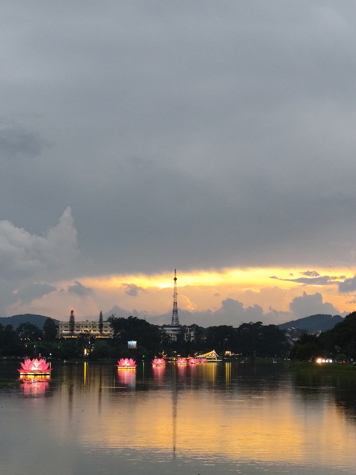 The image depicts a lake at sunset in Da Lat, Vietnam. In the foreground, the calm water reflects the sky, illuminated by a vibrant golden hue from the setting sun. Several illuminated, pink lotus flower-shaped lanterns float on the water, their reflections adding to the visual effect. Across the lake, a low-lying cityscape is visible, silhouetted against the sunset. A tall radio tower stands out against the sky, positioned slightly to the right of center. Mountains or hills line the horizon. The sky above is filled with heavy, gray clouds, hinting at an overcast or stormy evening. Vegetation, likely trees, is visible along the lake's edge, bordering the city. No people are directly visible.